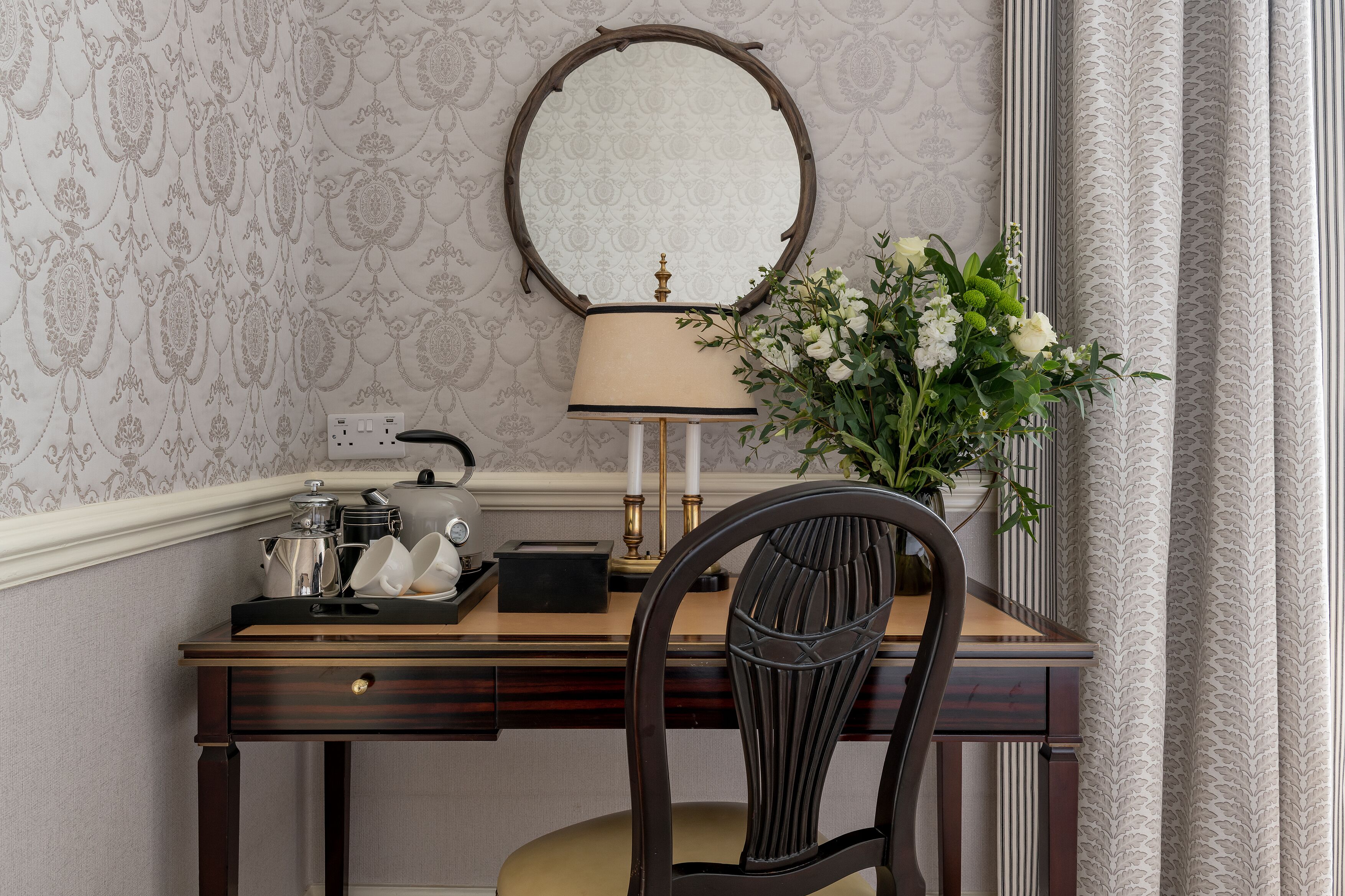 A wooden desk with a mirror, a lamp, and a vase of flowers.