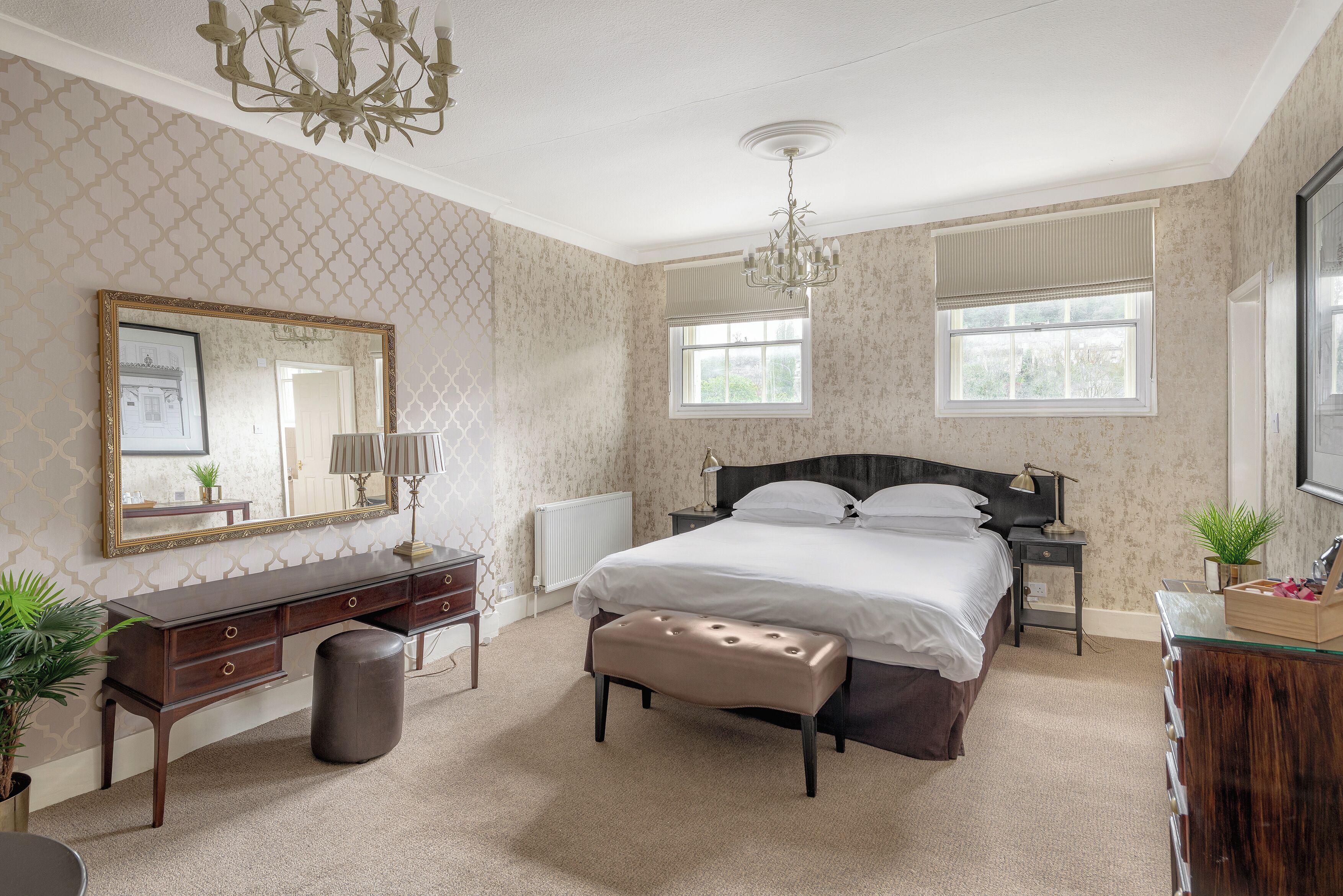 A bedroom with a large bed, a dresser, a mirror, a chandelier, and two windows.