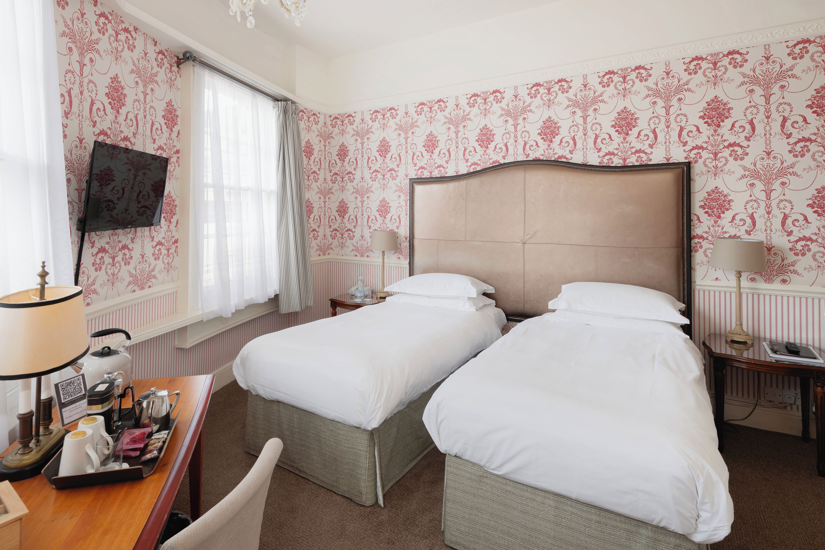A hotel room with two single beds, a desk with a chair, a television mounted on the wall, and a window with curtains.