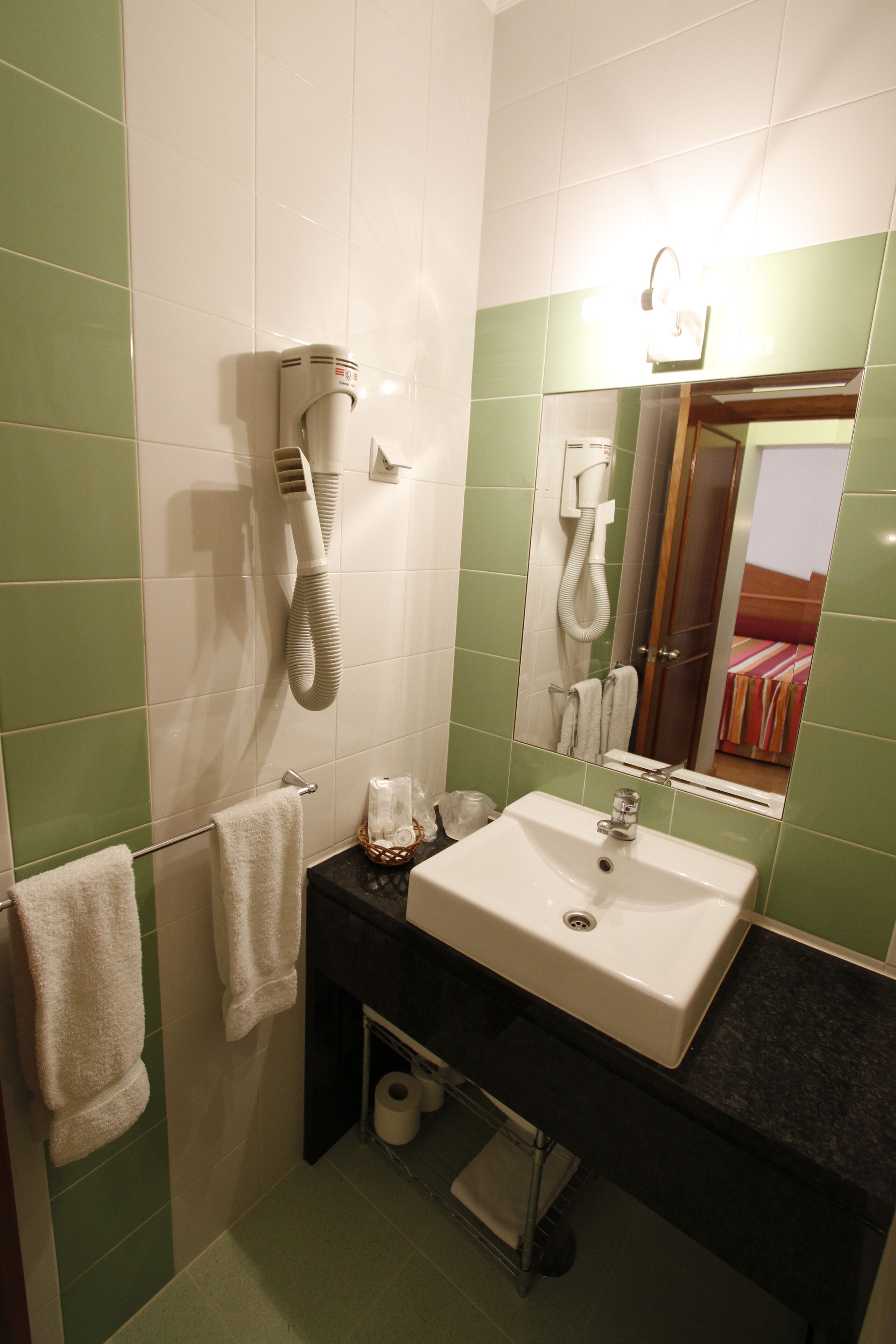 Un baño con un lavamanos blanco, baldosas verdes, un secador de pelo y un espejo que refleja una cama.