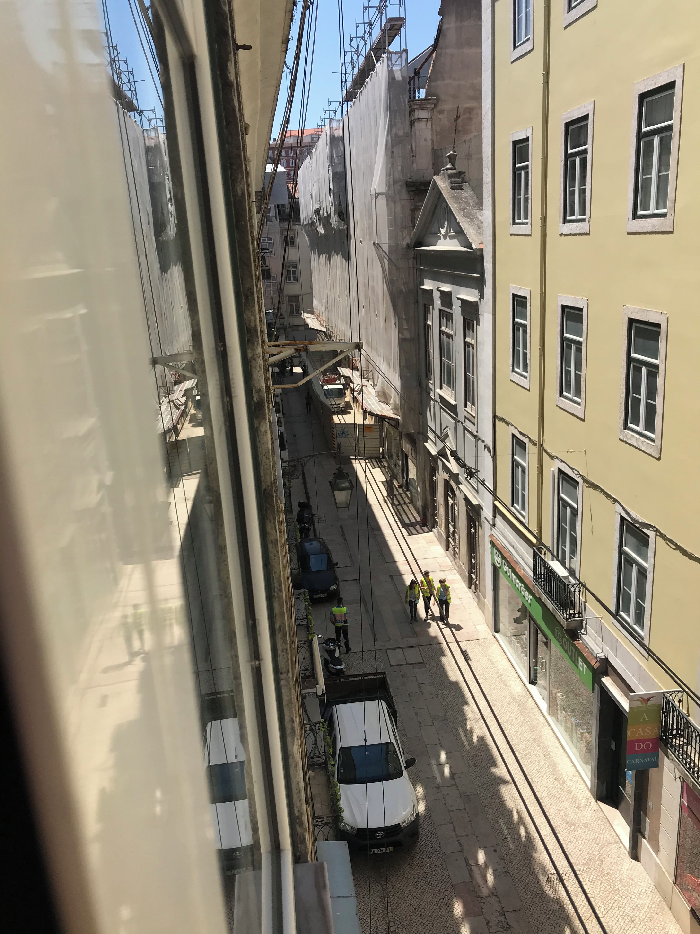 Ruas estreitas com edifícios em ambos os lados, uma carrinha branca estacionada na berma e alguns pedestres a caminhar.