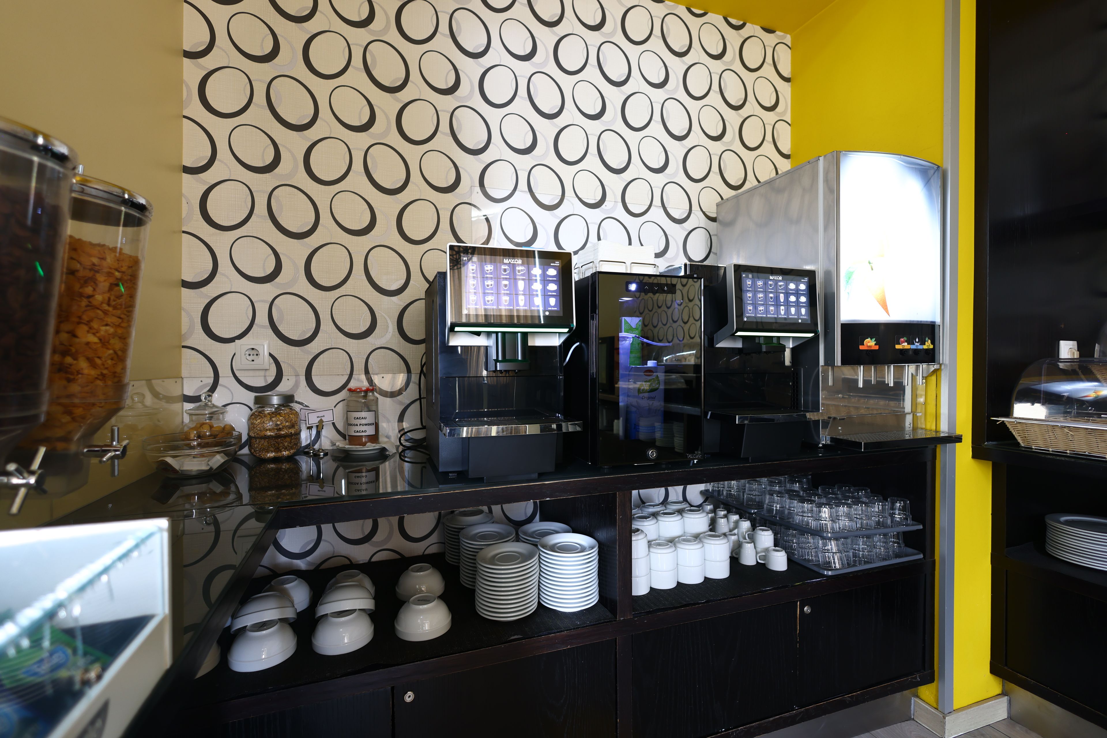 A coffee station with multiple coffee machines, a condiment dispenser, and a variety of coffee cups and saucers.