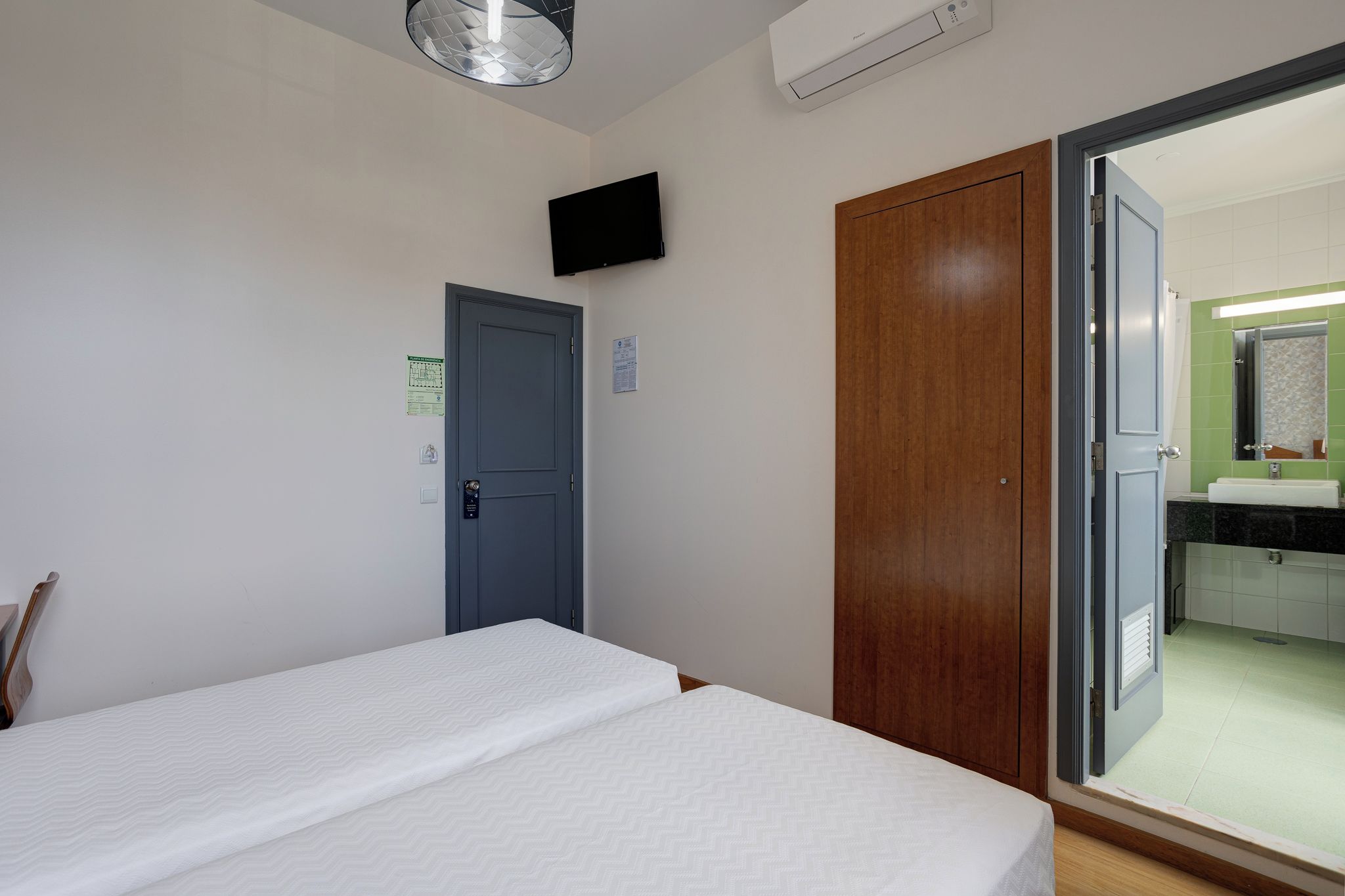 A hotel room with a bed, a wooden wardrobe, a door, and a bathroom visible through an open door.