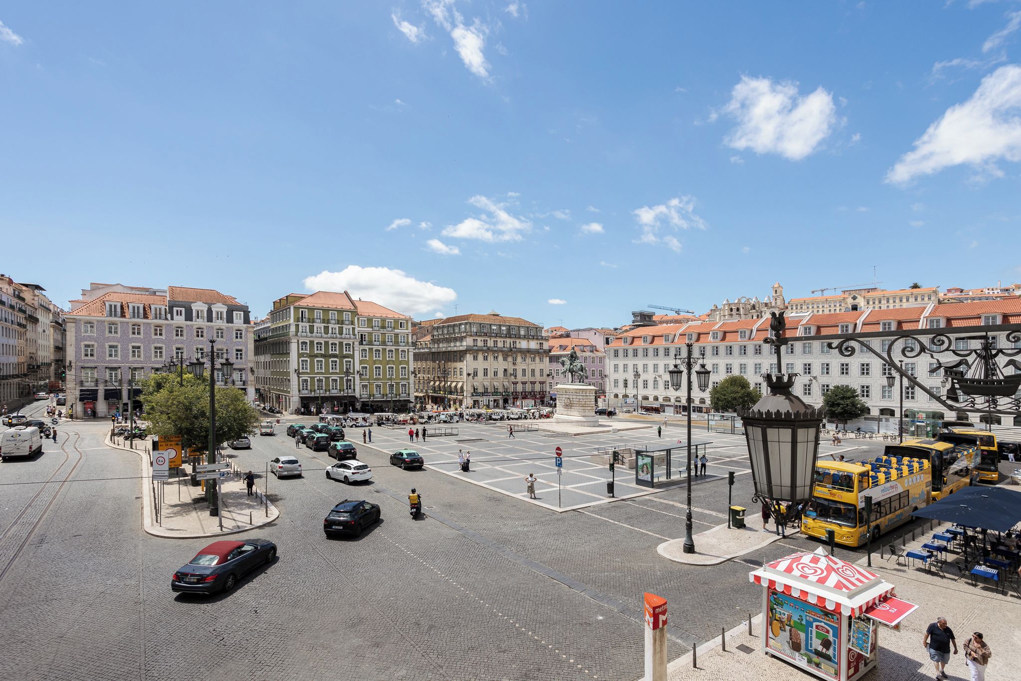 Une place de ville bordée de bâtiments historiques, ornée d’une statue et desservie par un arrêt de tramway.