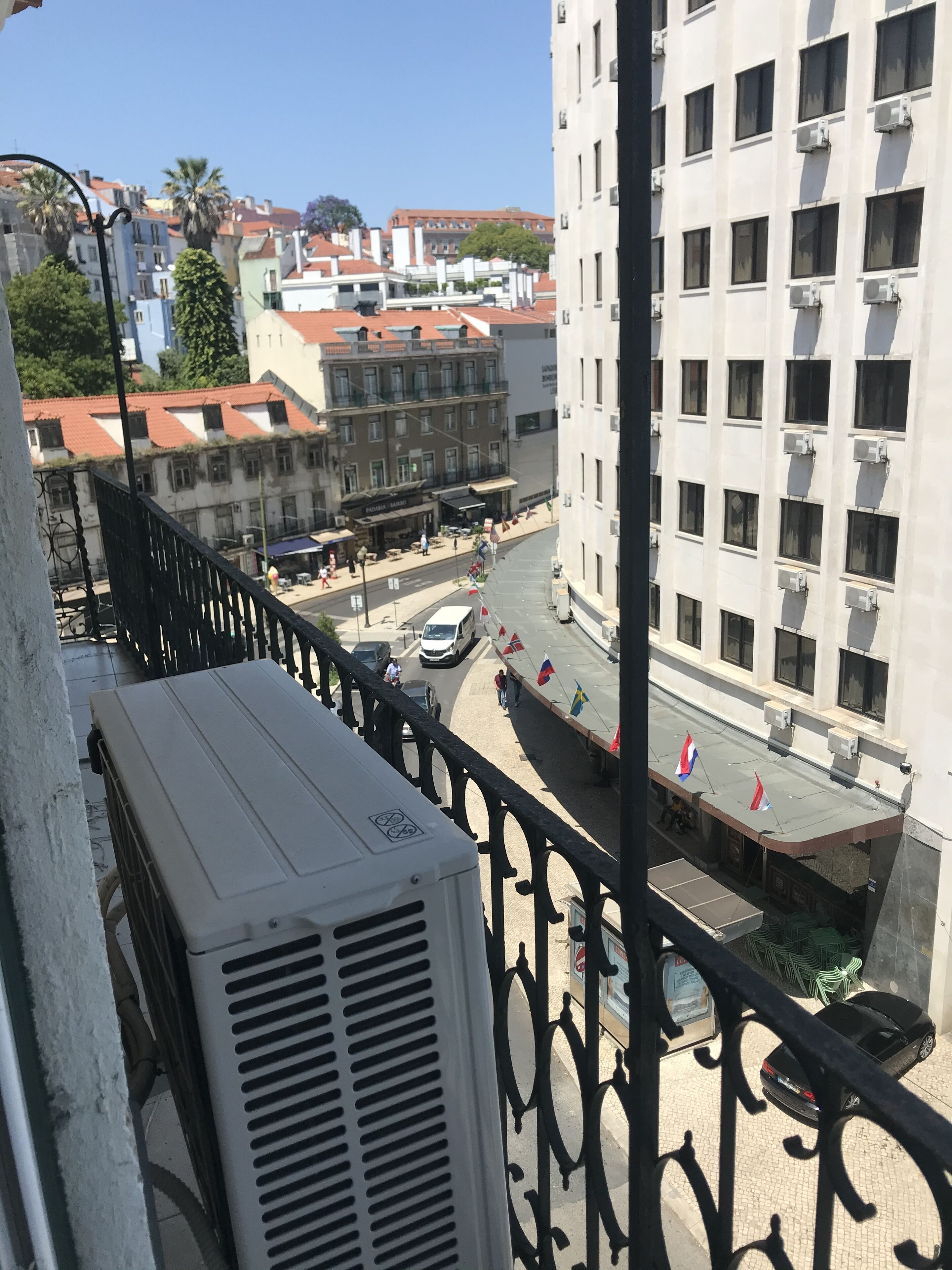 Un balcón con un equipo de aire acondicionado blanco, vistas a una calle con edificios, un coche estacionado y algunas personas caminando.