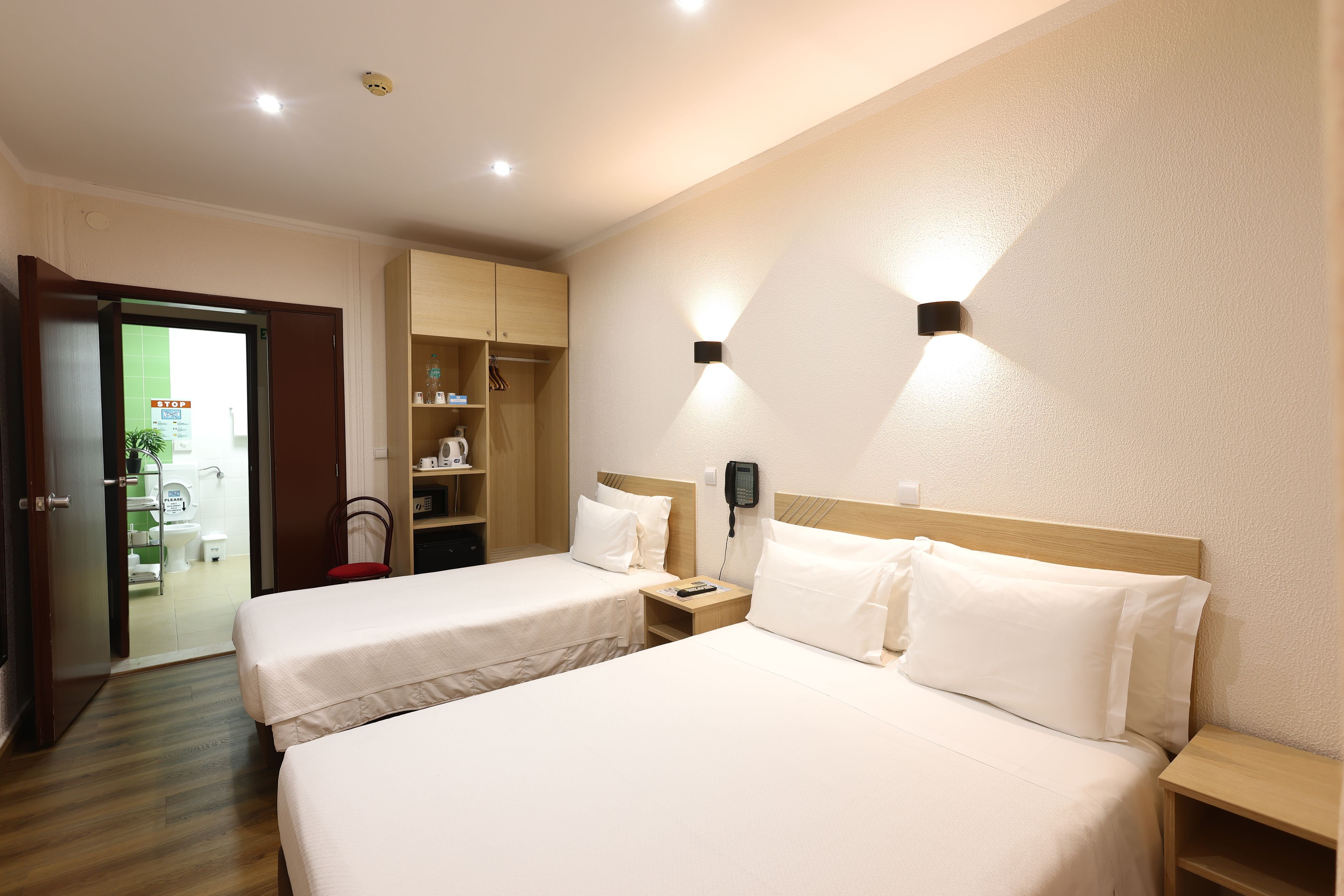 A hotel room with two beds, a wardrobe, a desk, a chair, and a bathroom visible through an open door.