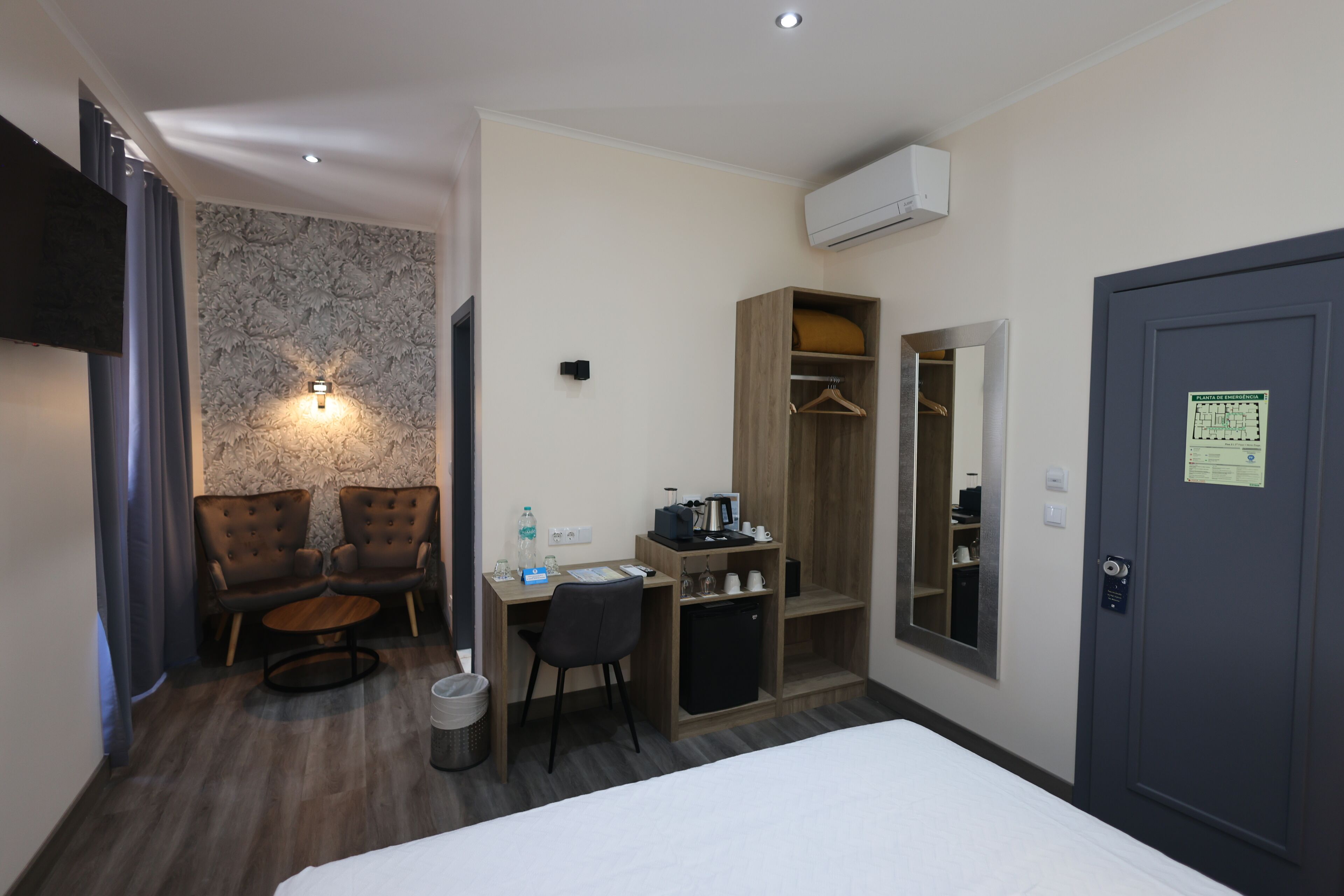 Una habitación de hotel con cama, escritorio con computadora, dos sillas, refrigerador pequeño, armario y equipo de aire acondicionado.