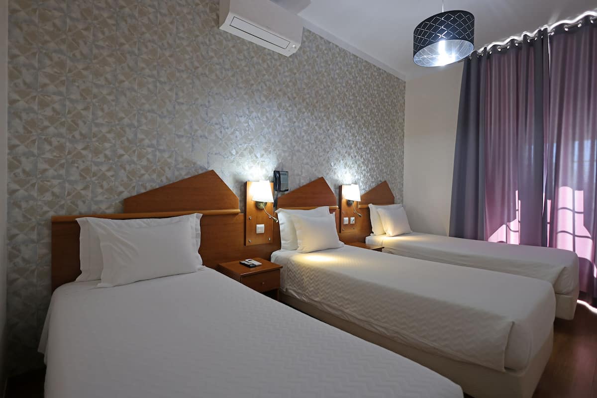 Een hotelkamer met drie bedden, een houten hoofdeinde, wandlampen en een groot raam met gordijnen.