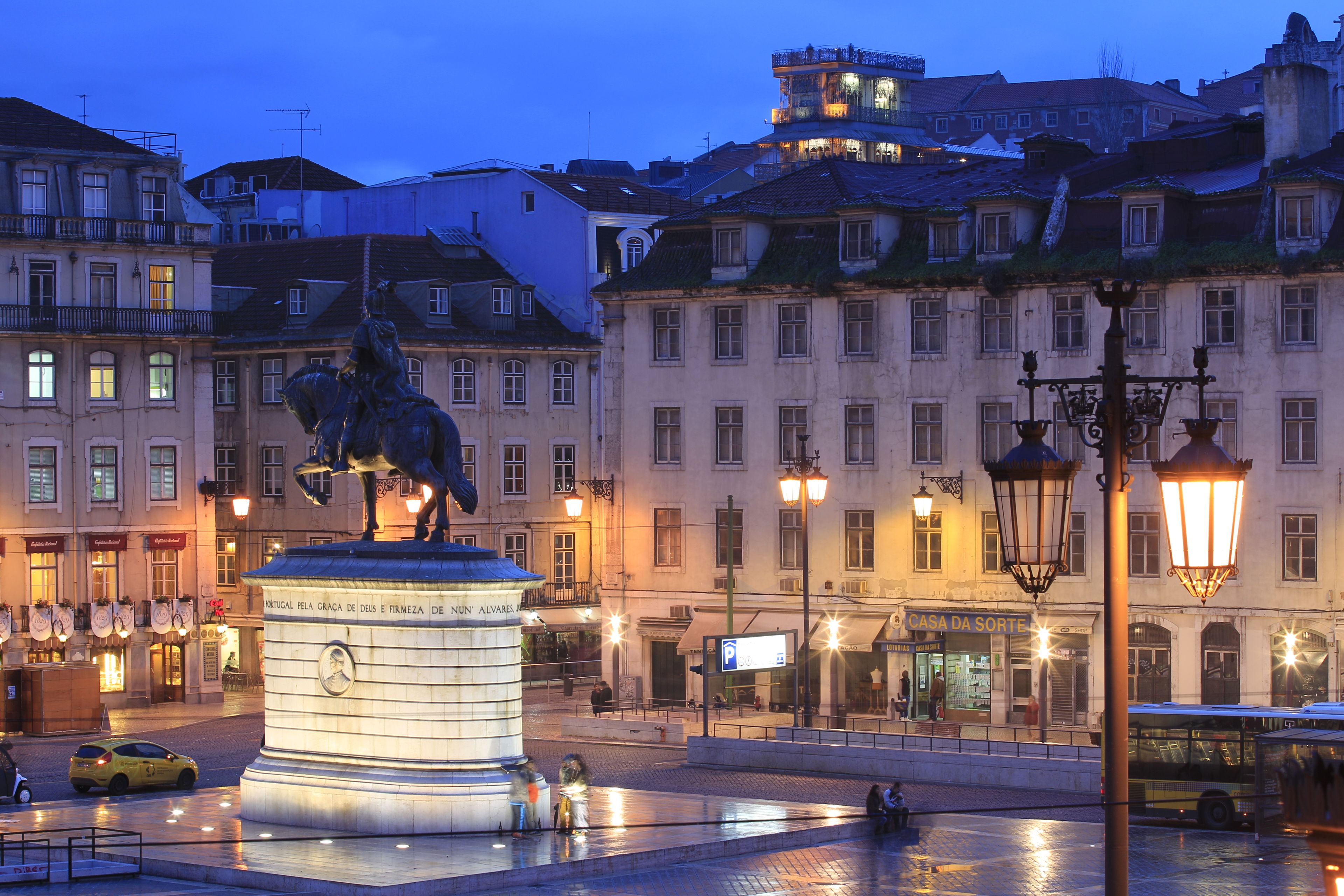 Uma praça histórica com um monumento central, edifícios ao redor e um ônibus ao fundo.