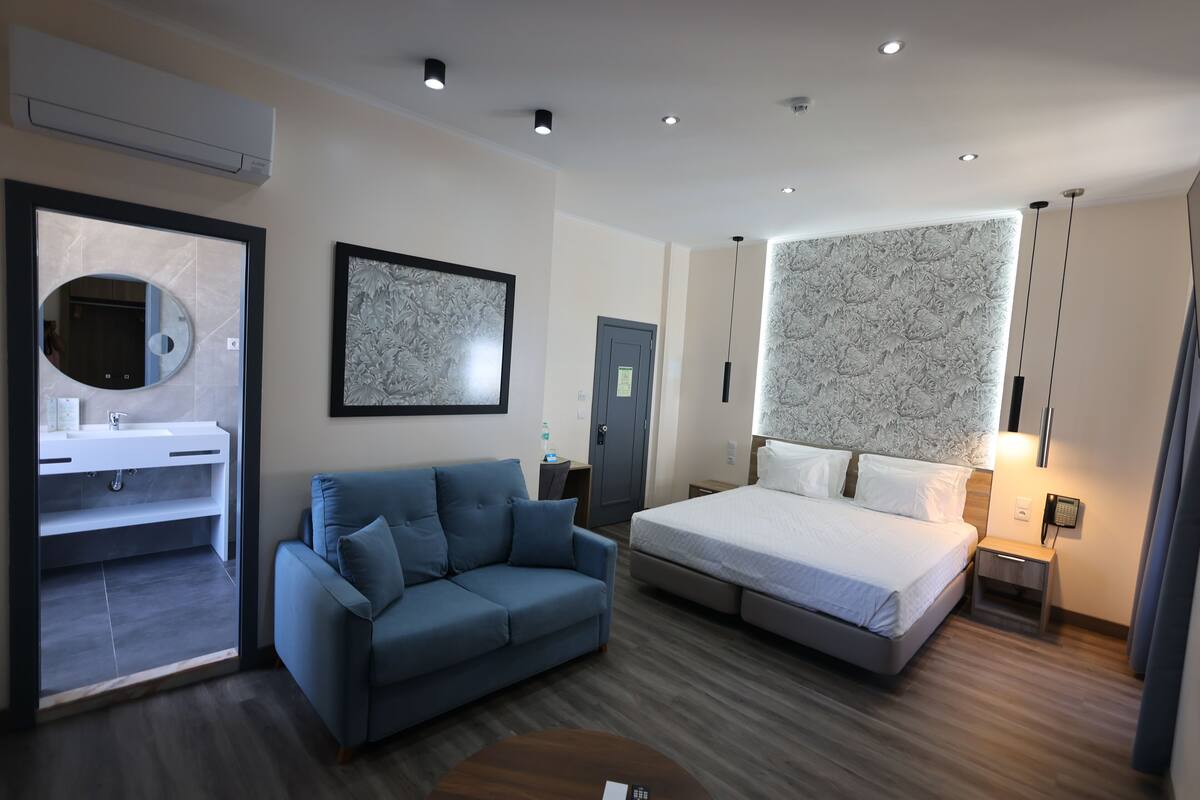 Een moderne hotelkamer met een bed, een bank, een bureau, een spiegel en een badkamer, zichtbaar door een geopende deur.