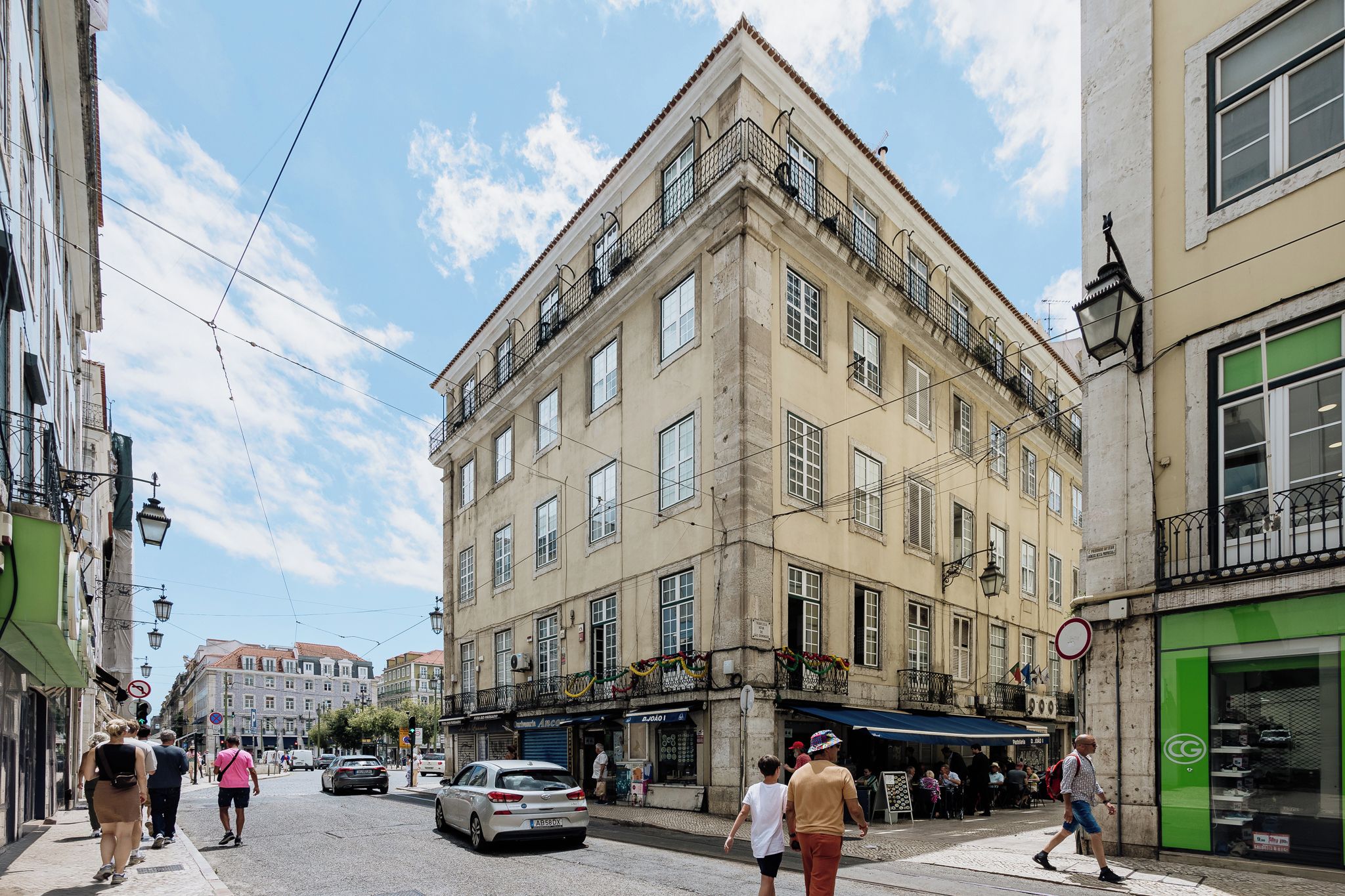 Une vue de rue avec un immeuble à plusieurs étages doté de balcons, un magasin au rez-de-chaussée et des piétons.