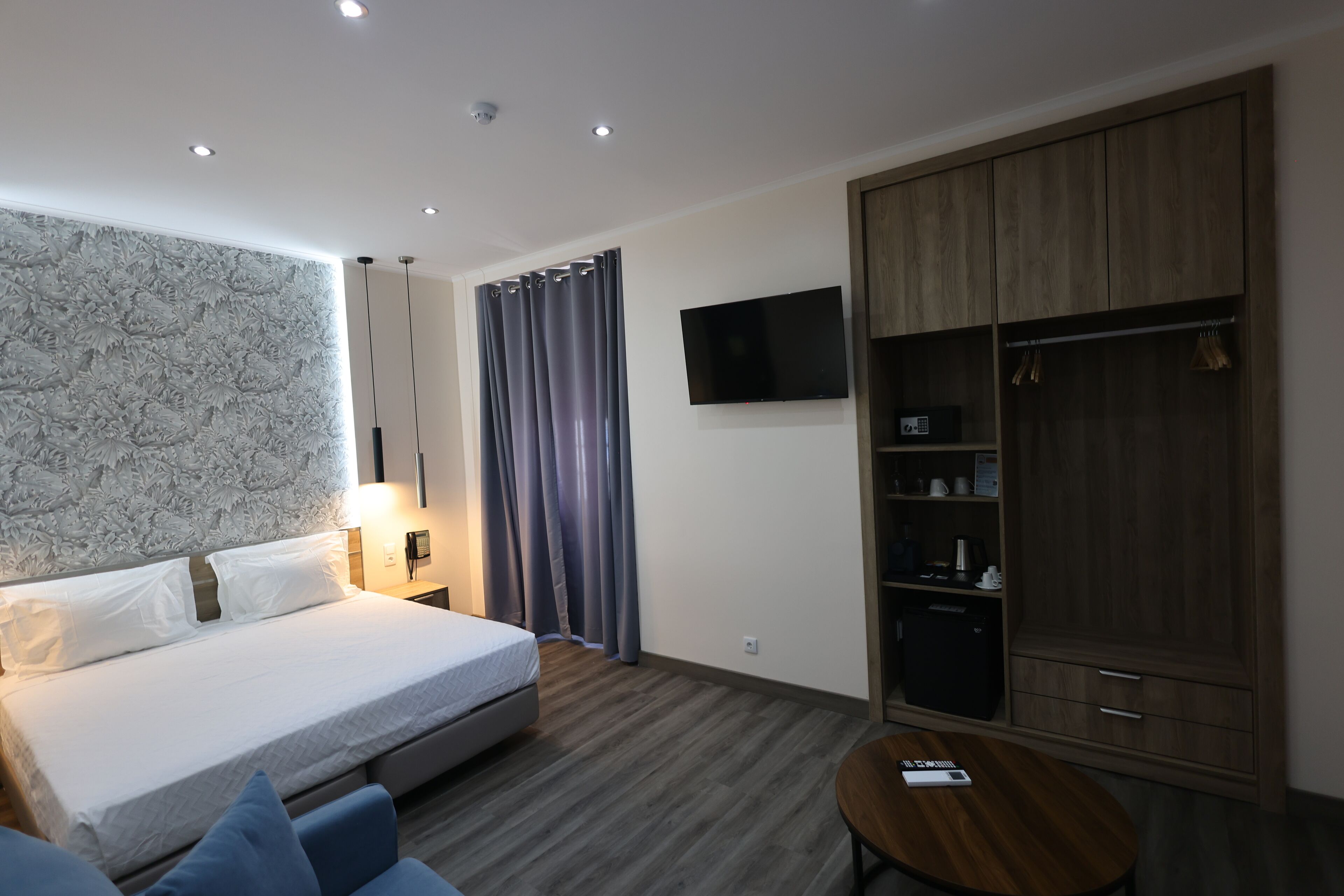 A modern hotel room with a large bed, a wooden wardrobe, a TV mounted on the wall, and a small table.