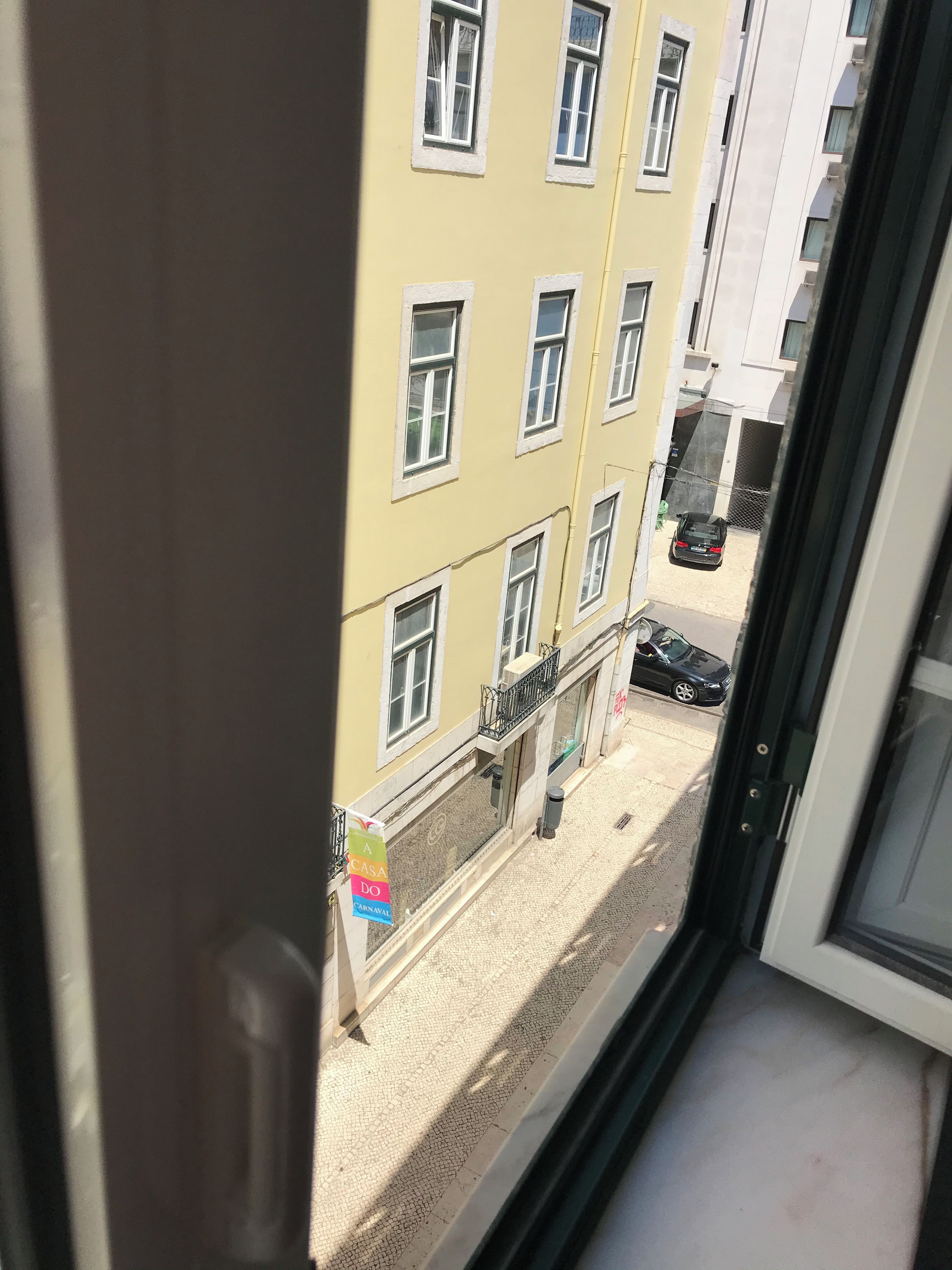 Udsigt fra et vindue over en gade med en gul bygning, en sort bil parkeret på gaden, og et skilt med farverig tekst.