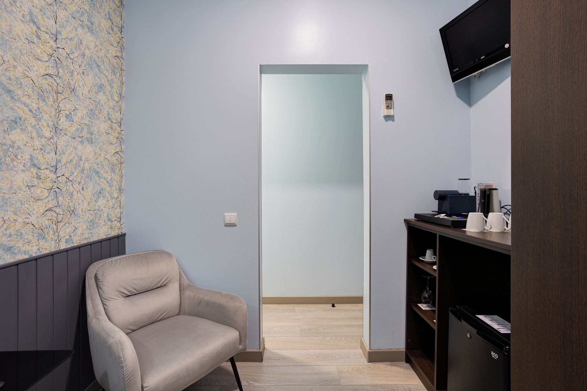 Una habitación con una silla, una cafetera, un televisor de pantalla plana y una puerta que da a otra habitación.