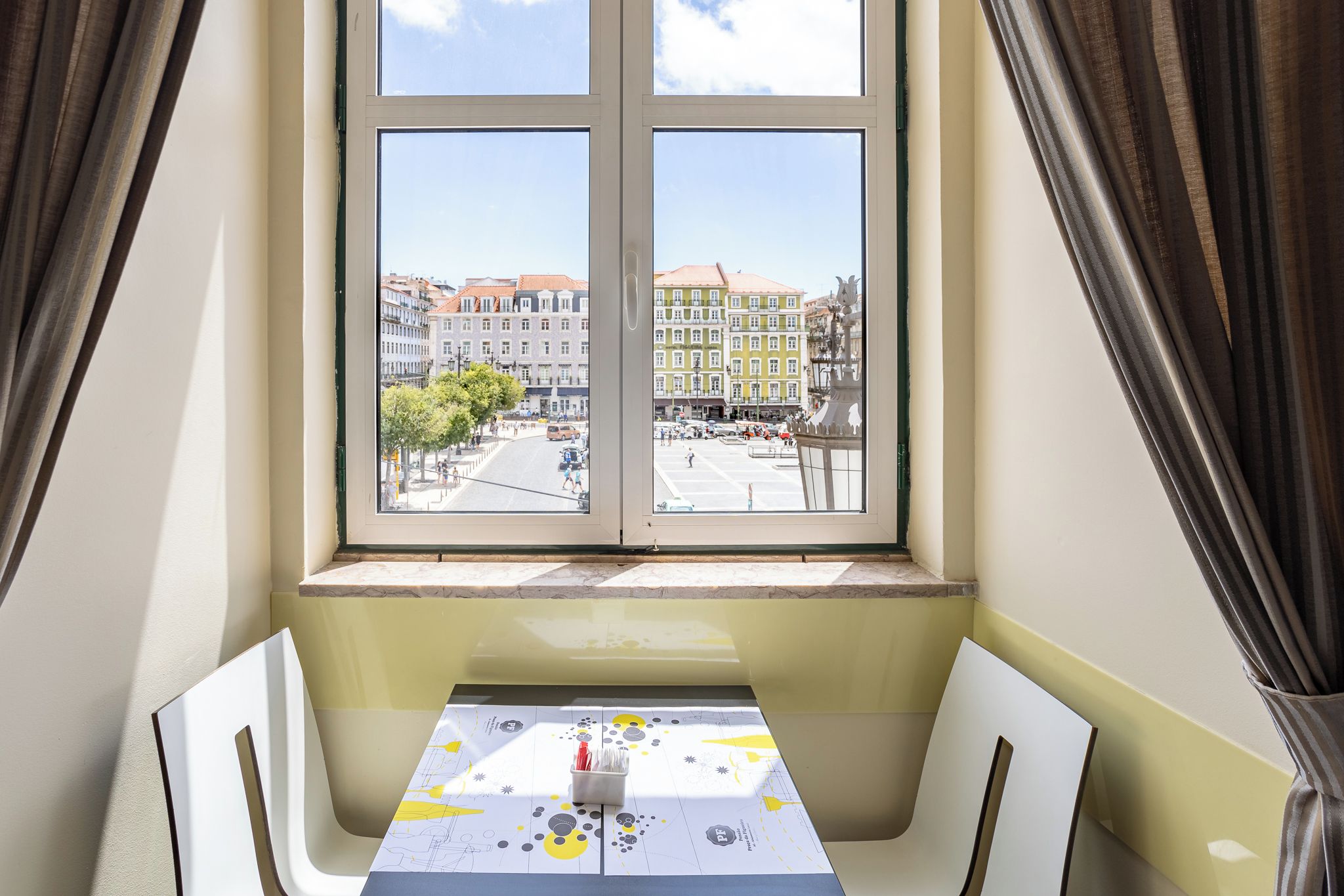 Una habitación con una mesa, sillas, una ventana que ofrece vistas a edificios y una cortina.