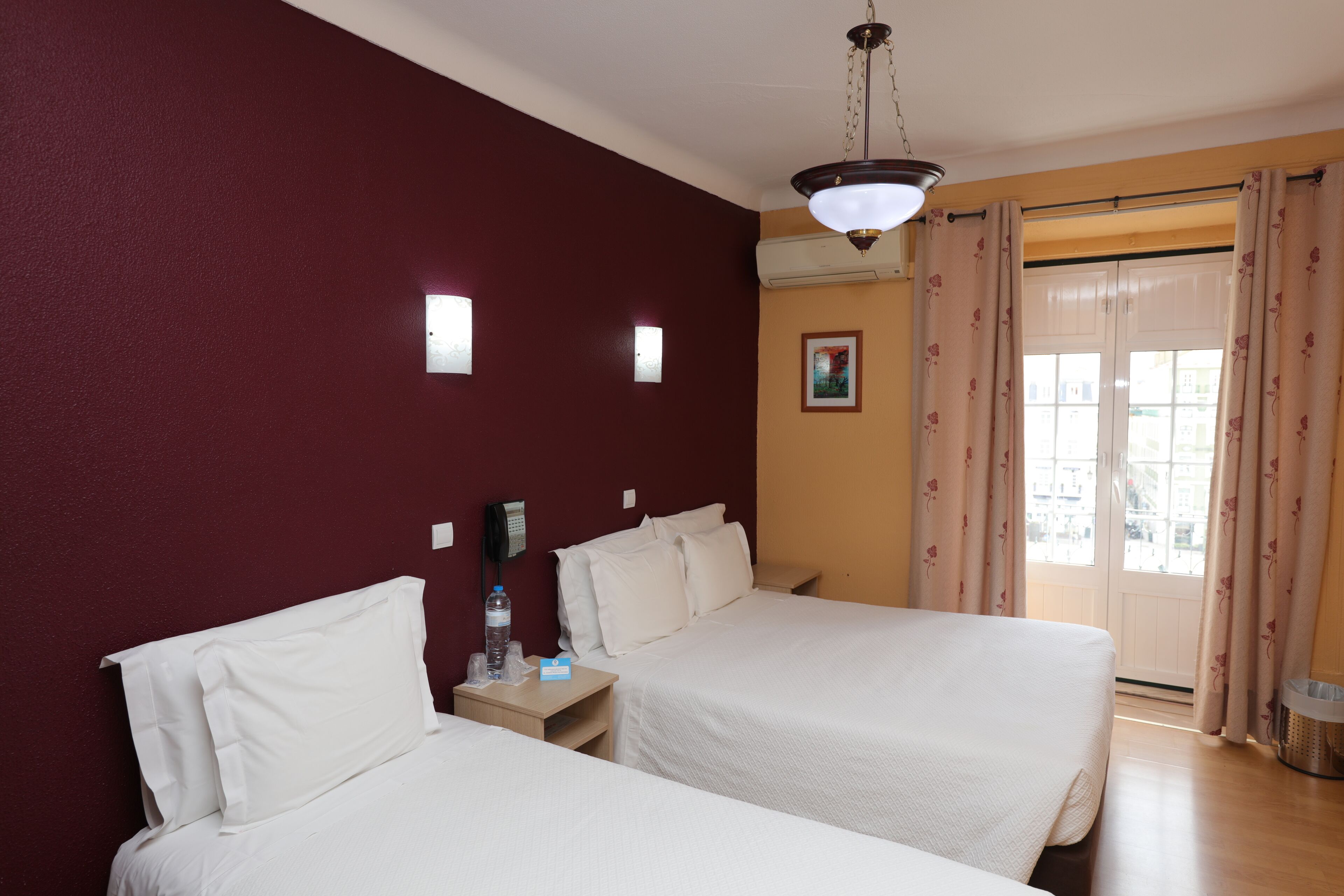 Quarto de hotel com duas camas, uma luminária no teto, ar-condicionado fixado na parede, uma janela com cortinas e uma pequena imagem na parede.