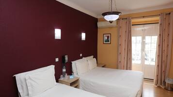 A hotel room with two beds, a ceiling light, a wall-mounted air conditioner, a window with curtains, and a small picture on the wall.