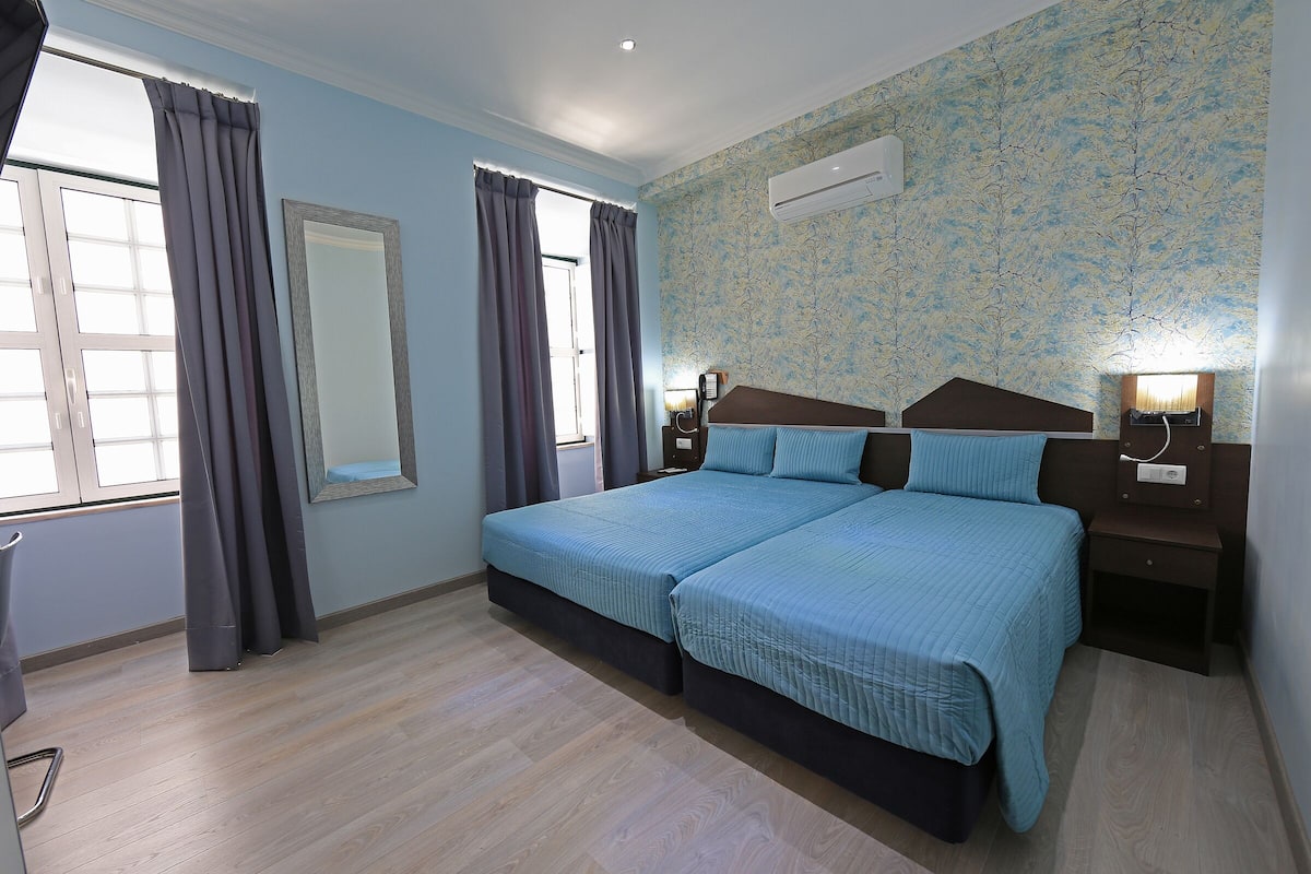 Een slaapkamer met twee bedden, een groot raam met gordijnen, een spiegel en een wandairconditioner.