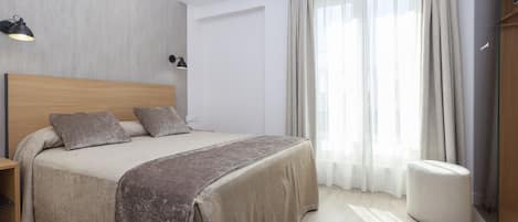 A modern bedroom with a large bed, bedside lamps, a wooden headboard, a bedside table, and a large window with curtains.
