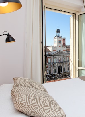 Habitación de hotel con una cama, un cojín estampado, una lámpara de escritorio negra y vistas a un edificio histórico con una torre de reloj.