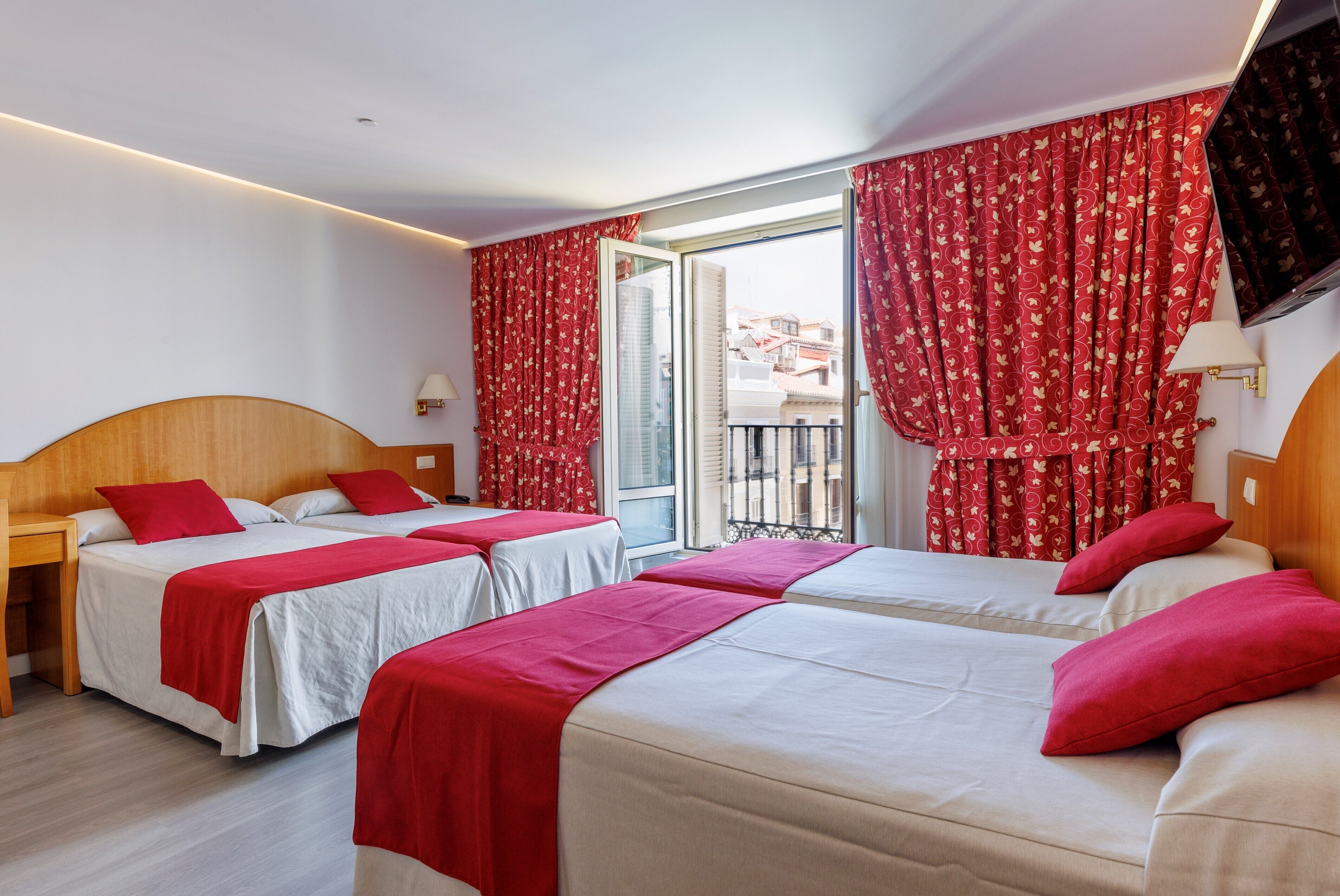 Camera d'albergo con due letti, tende rosse, una TV e un balcone con vista.