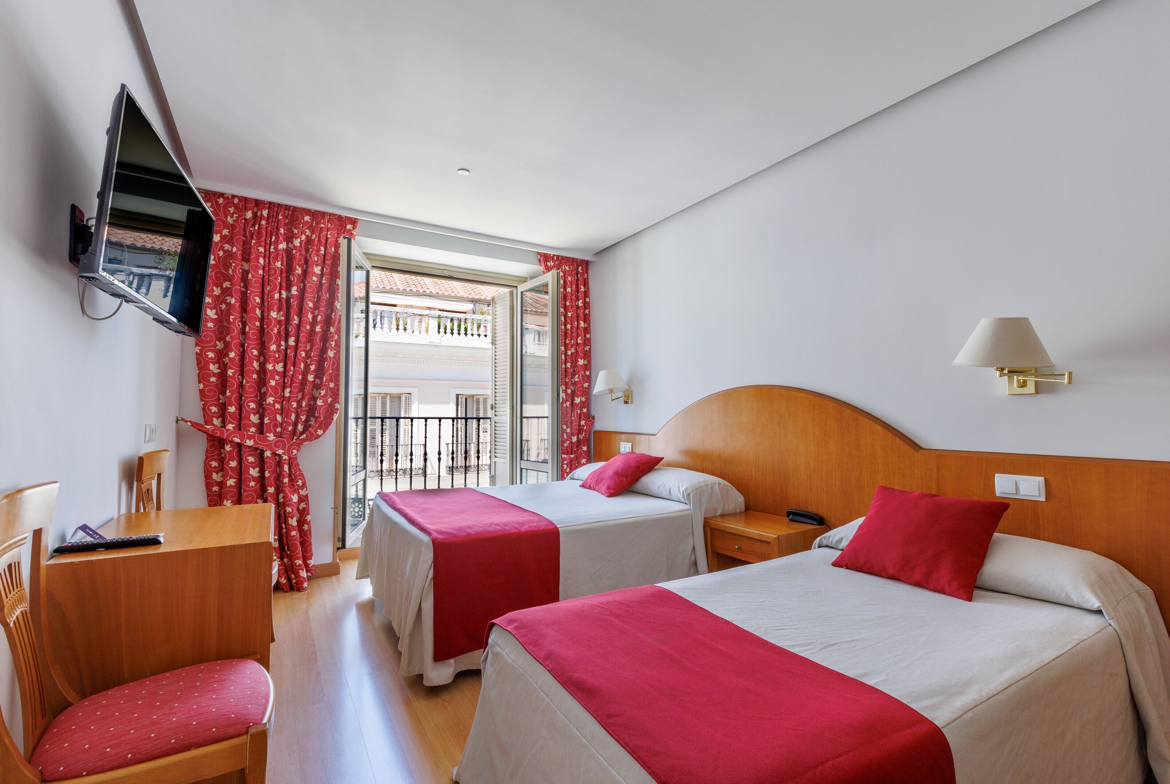 Habitación de hotel con dos camas, un televisor montado en la pared, un escritorio con una silla y una ventana con cortinas rojas.