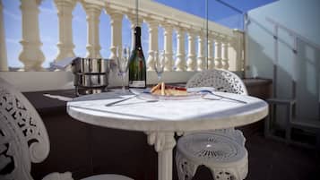 Une table ronde avec une bouteille de champagne, un verre et une part de gâteau, le tout posé sur un balcon agrémenté de chaises blanches et d’une balustrade.