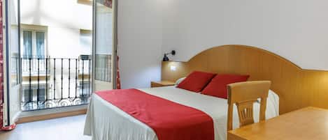A hotel room with a bed, a wooden headboard, a nightstand, a desk, and a chair. There is a balcony with a railing and a view of the outside.