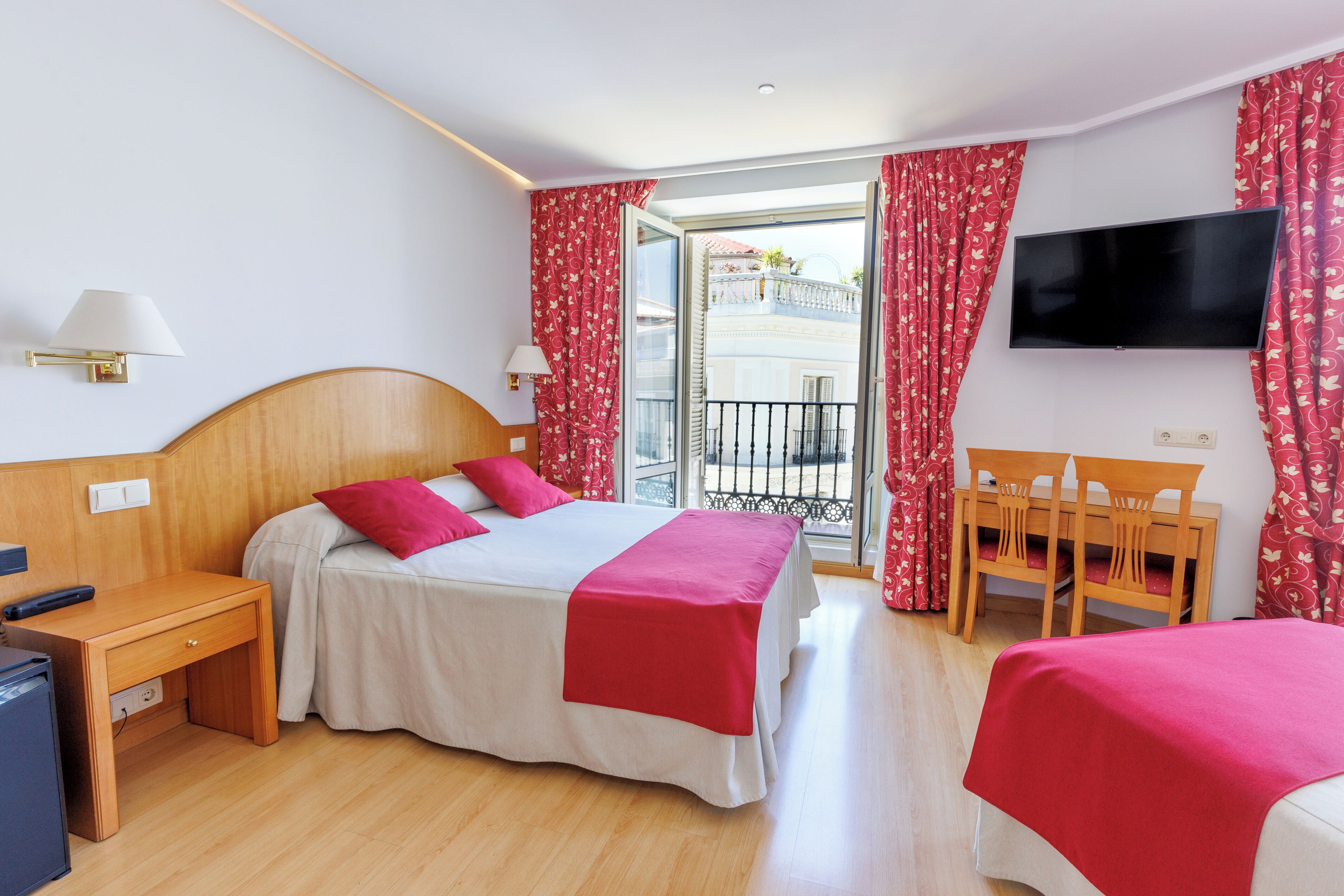 A hotel room with a bed, a desk, a chair, a TV, and a balcony with red curtains.