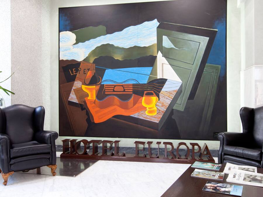 Un vestíbulo con dos sillones de cuero negro, un cuadro abstracto grande y un mostrador de recepción con la señalización 'HOTEL EUROPA'.
