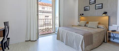 A hotel room with a bed, a desk, and a chair. There is a balcony with a view of buildings.