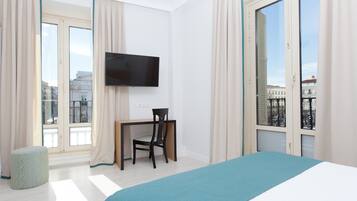 Una habitación de hotel con una cama, un escritorio, una silla, un televisor y ventanas amplias con cortinas.