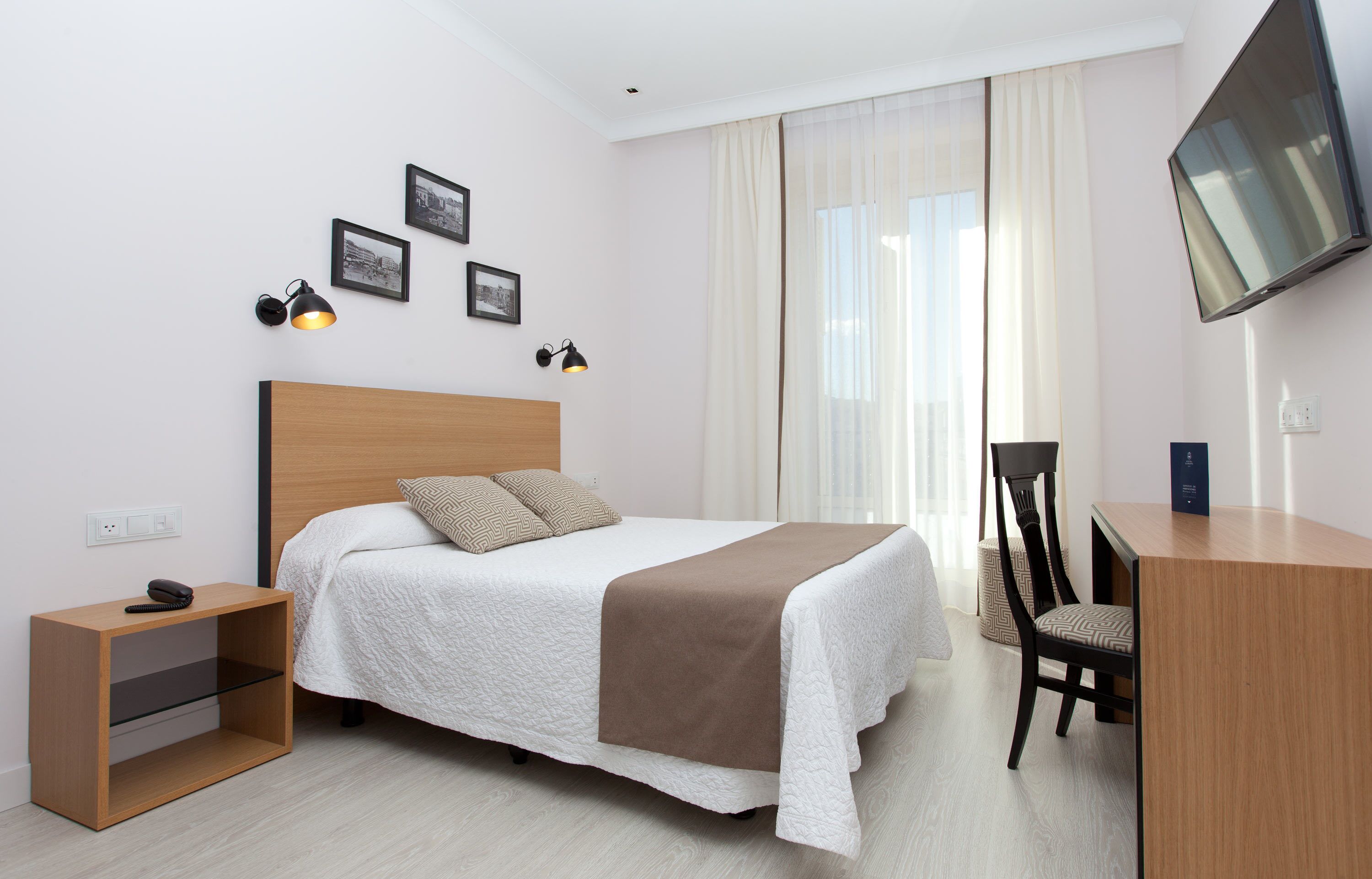 Ein ordentlich eingerichtetes Hotelzimmer mit Bett, Nachttisch, Sessel und einem an der Wand befestigten Fernseher.