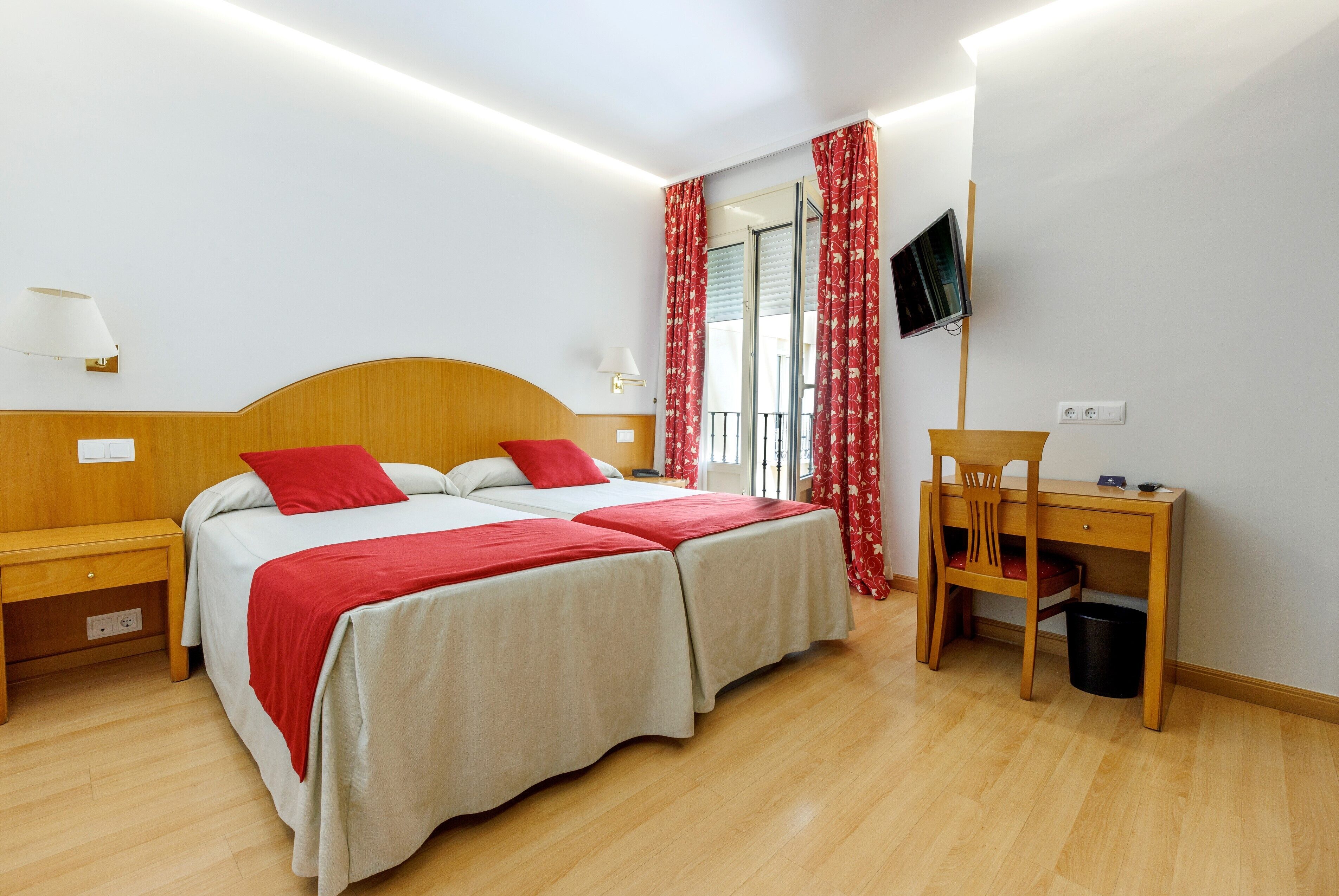 A hotel room with two beds, a desk, a chair, a TV, and a window with curtains.