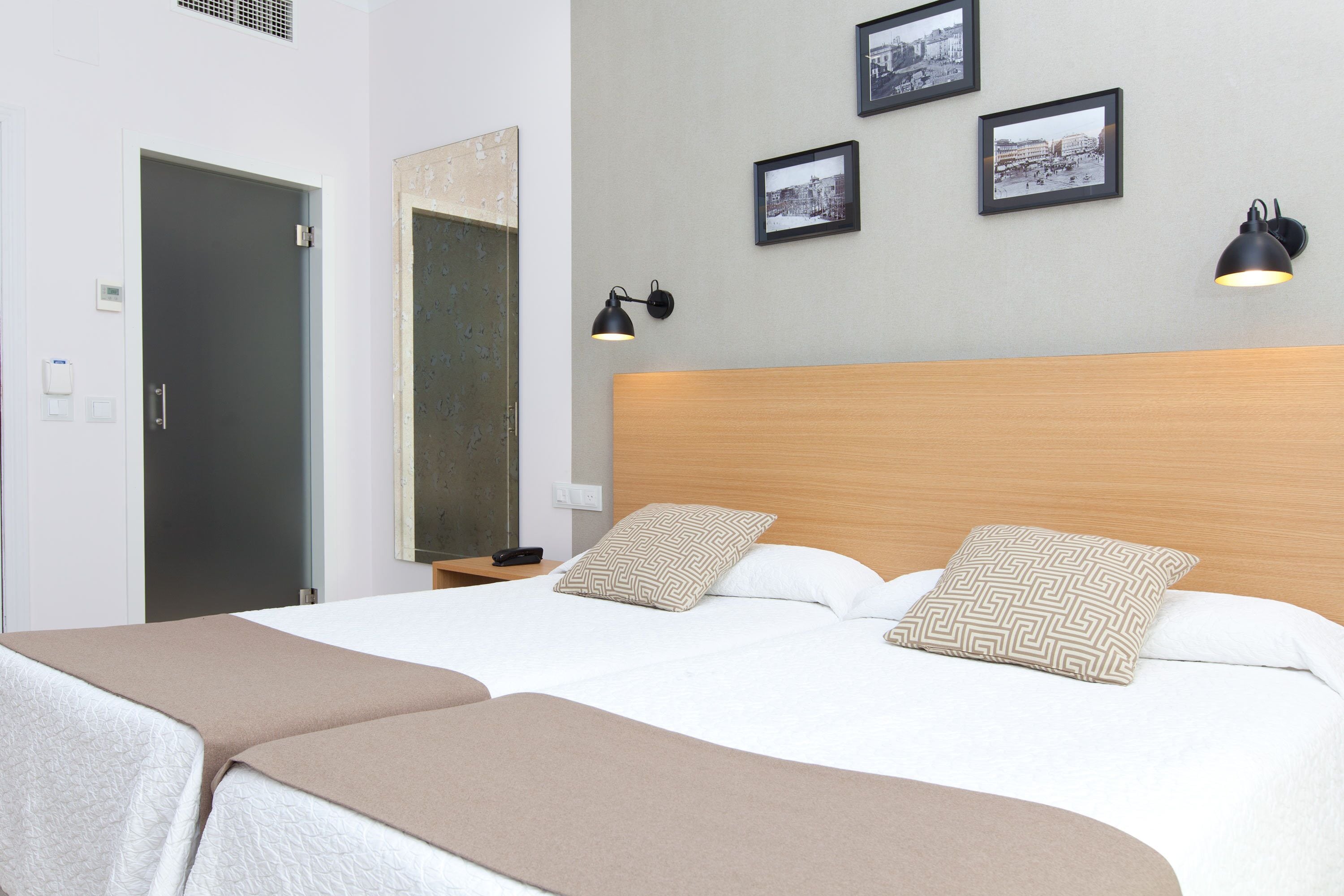A double bed with two pillows, a wooden headboard, and a beige throw blanket. There are two wall-mounted lamps and three framed pictures on the wall.