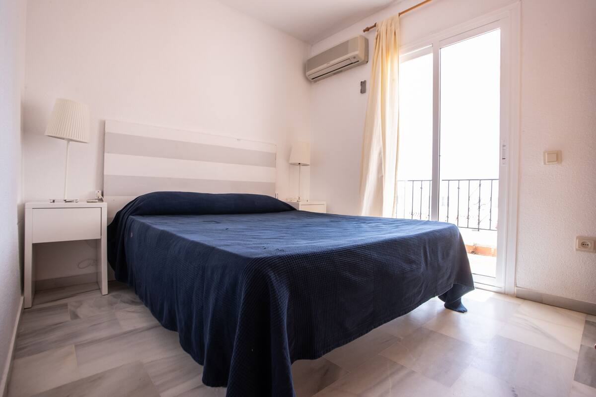Un dormitorio con cama, mesita de noche, lámpara y ventana con cortinas.