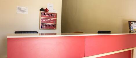 A reception desk with a red counter and a wooden desk to the left. There is a bulletin board with notices on the wall behind the desk.