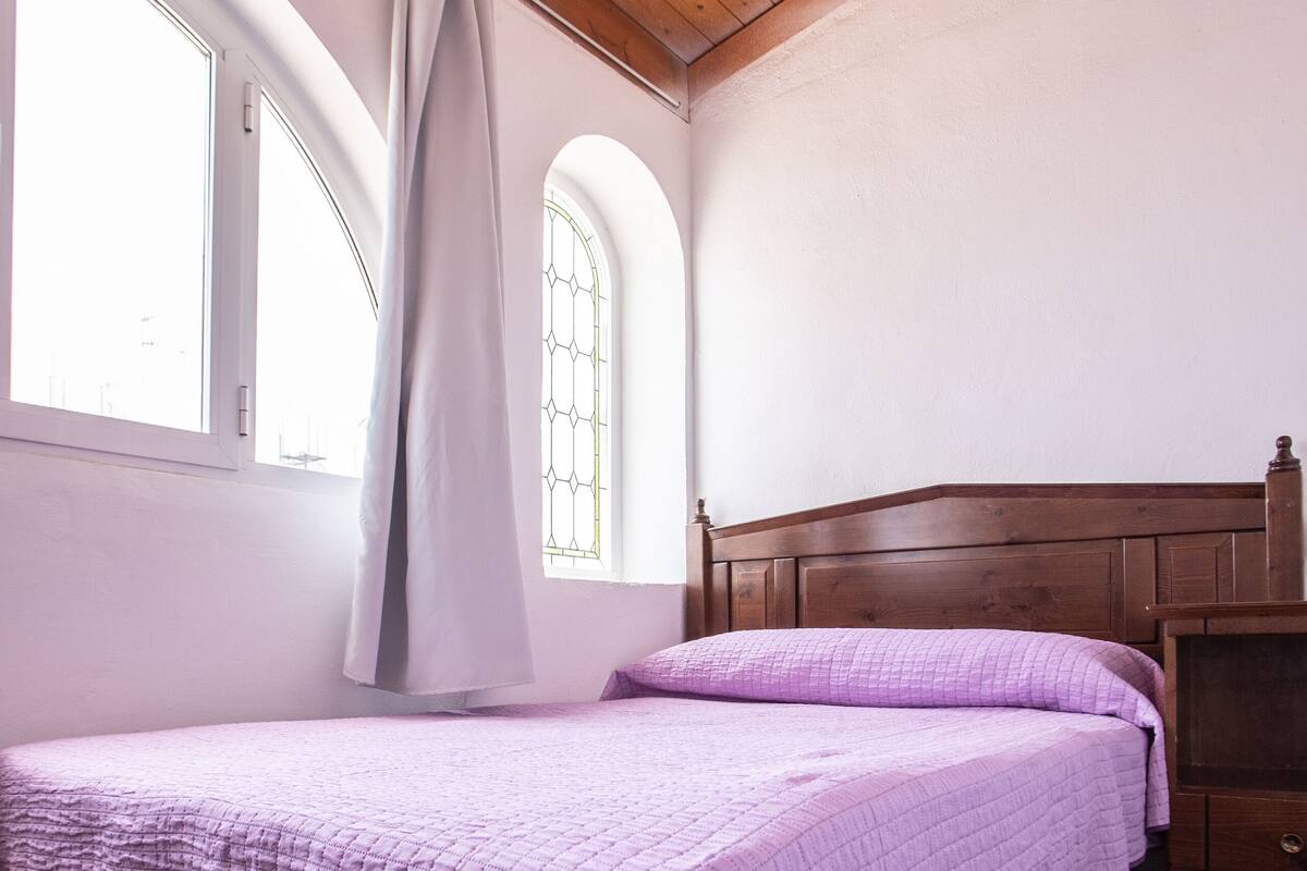 Un dormitorio con techo de madera, una cama con colcha morada, una mesita de noche y una ventana con cortina.