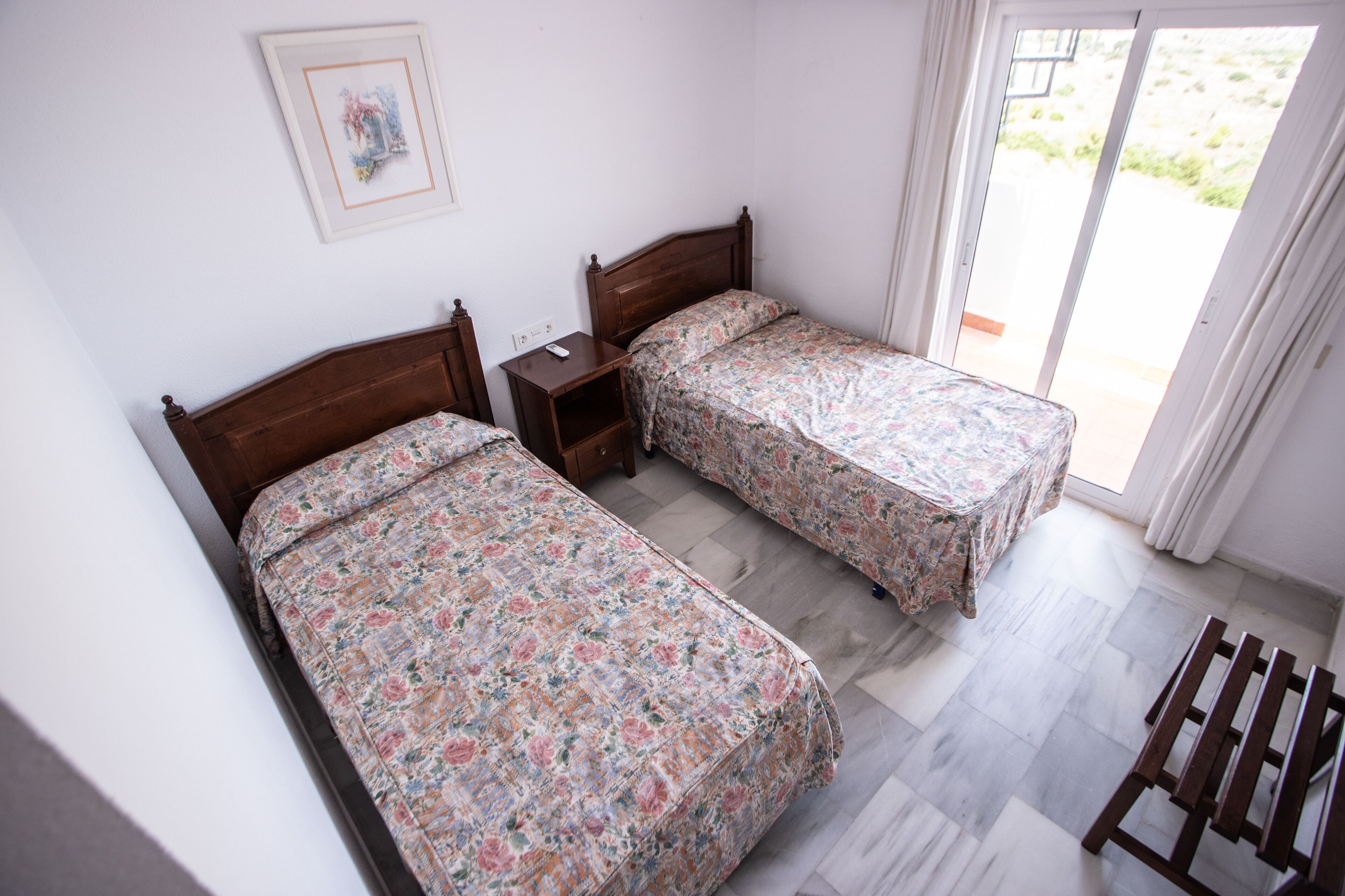 A bedroom with two single beds, a nightstand, a wooden bench, and a sliding glass door leading to an outdoor area.
