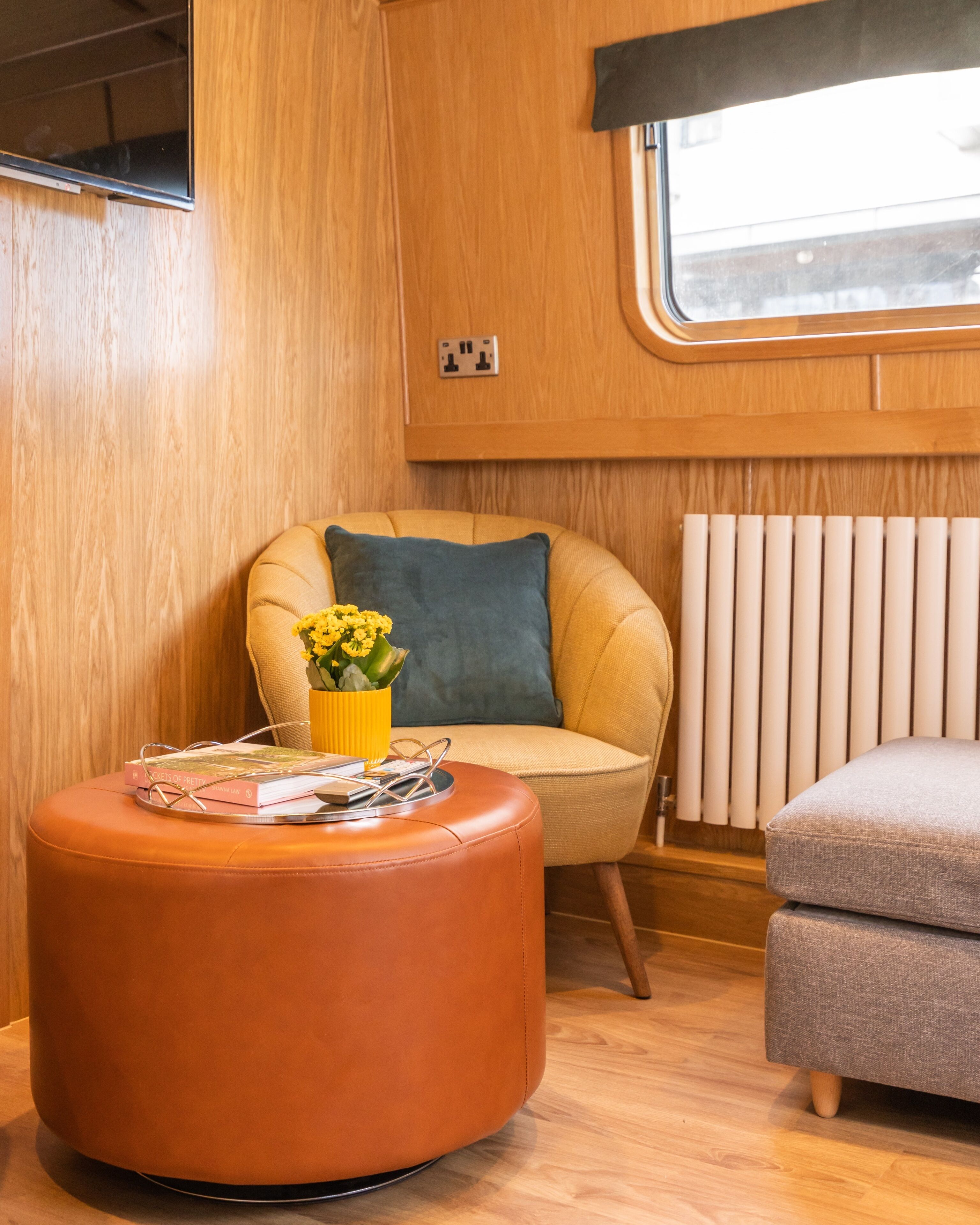 Un interior acogedor con un revestimiento de madera, una ventana con persianas, un sillón con cojín azul, un banquillo naranja redondo con libros y un jarrón amarillo, y un sofá gris.