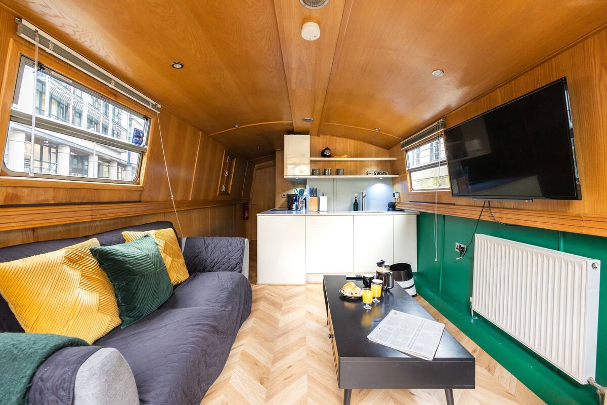 A compact interior with a sofa, a small table with breakfast items, a kitchen area, and a TV.