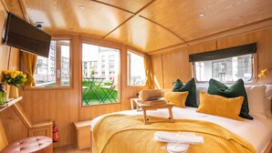 A modern hotel room with a wooden interior, a bed with yellow bedding, a small table with a book, and a TV mounted on the wall. - The Four Sisters Boatel (Edinburgh)
