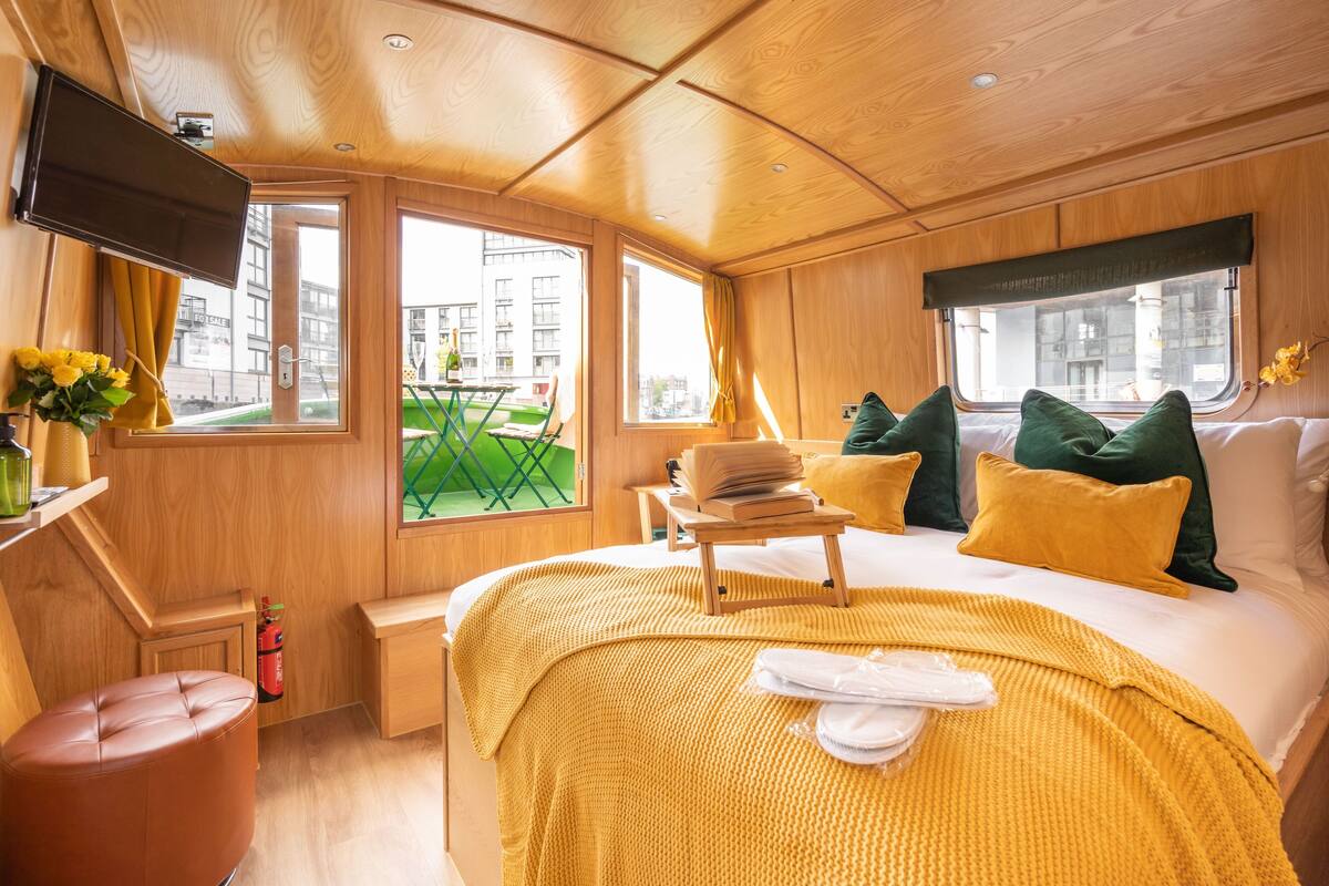 A modern hotel room with a wooden interior, a bed with yellow bedding, a small table with a book, and a TV mounted on the wall.