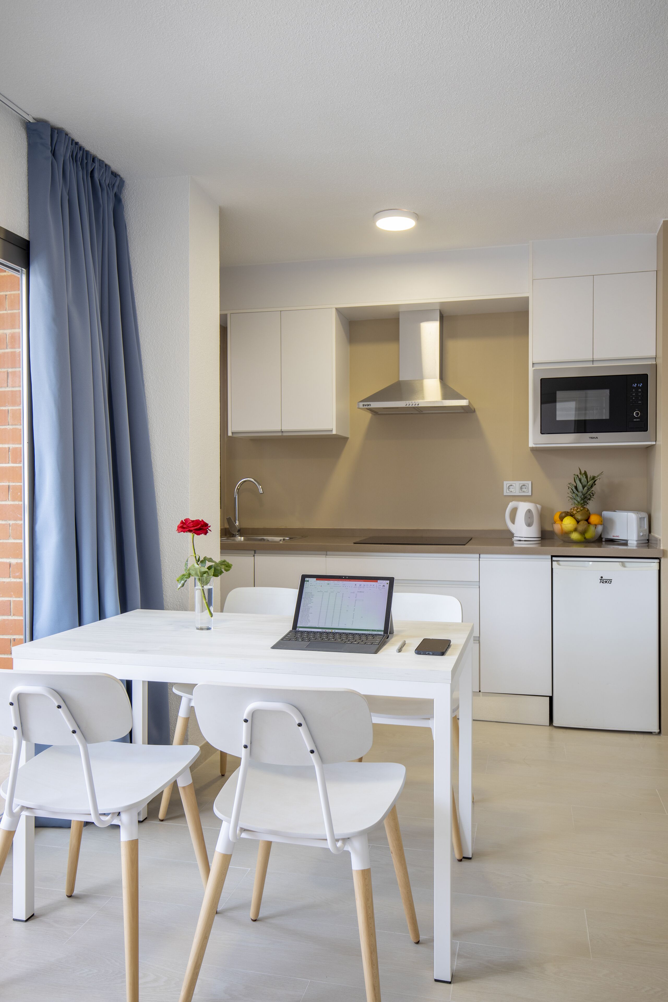 A modern kitchen with white cabinets, a microwave, and a sink. There is a laptop on the table, a vase with a red flower, and a pineapple on the counter.