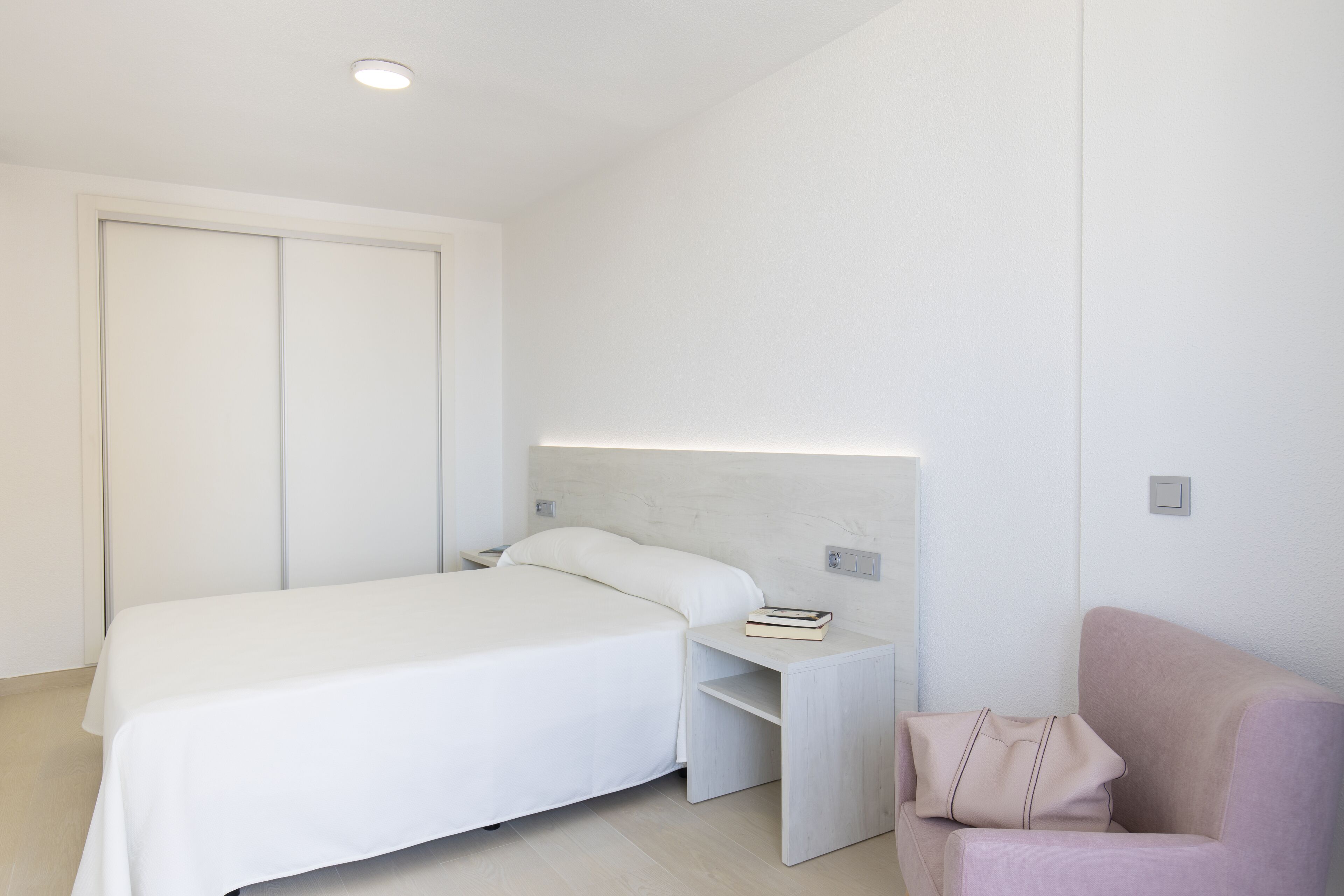 Ett modernt hotellrum med en stor säng, ett nattduksbord med en bok, en sänglampa och en rosa stol.