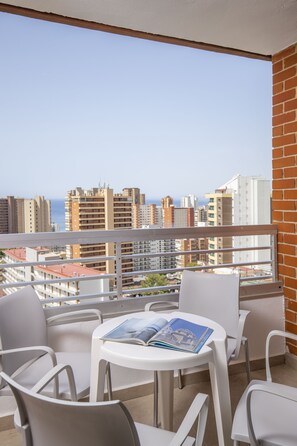 Een balkon met witte stoelen en een tafel, met uitzicht op een stadsgezicht met gebouwen en een heldere lucht.