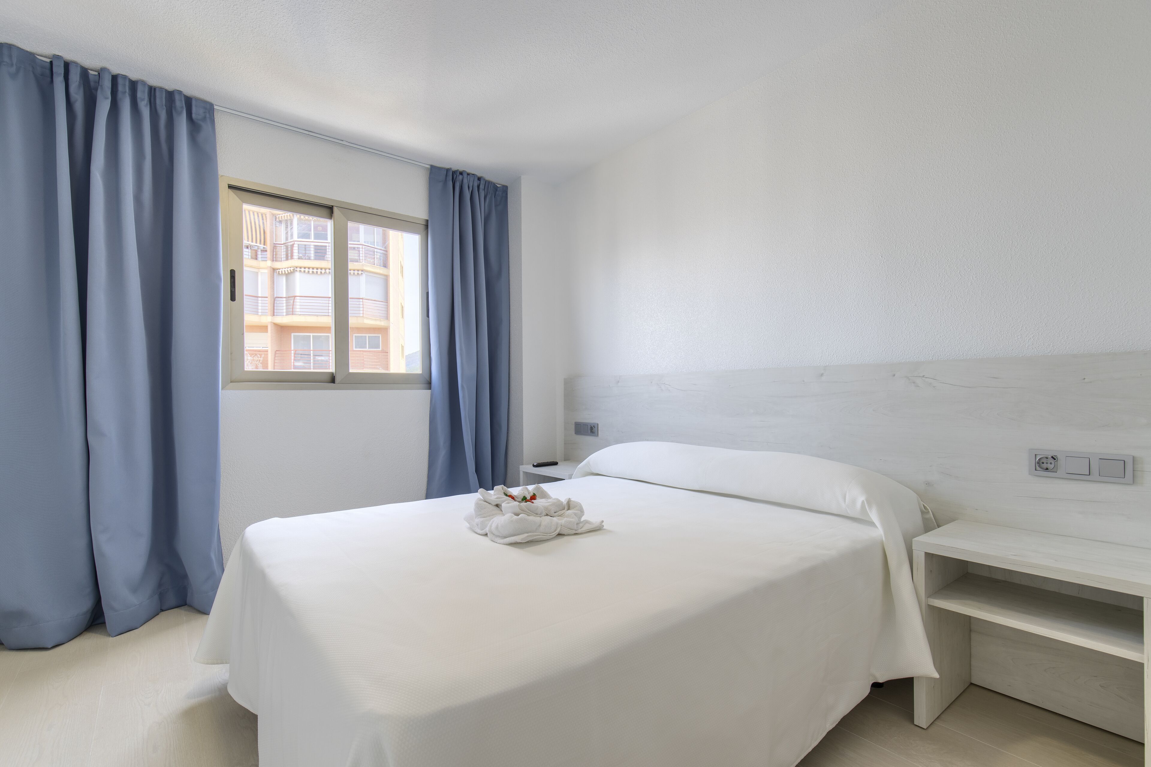 Quarto de hotel com cama, mesa de cabeceira, janela com cortinas e vista para um edifício.