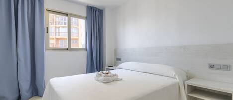 A hotel room with a bed, a bedside table, a window with curtains, and a view of a building outside.