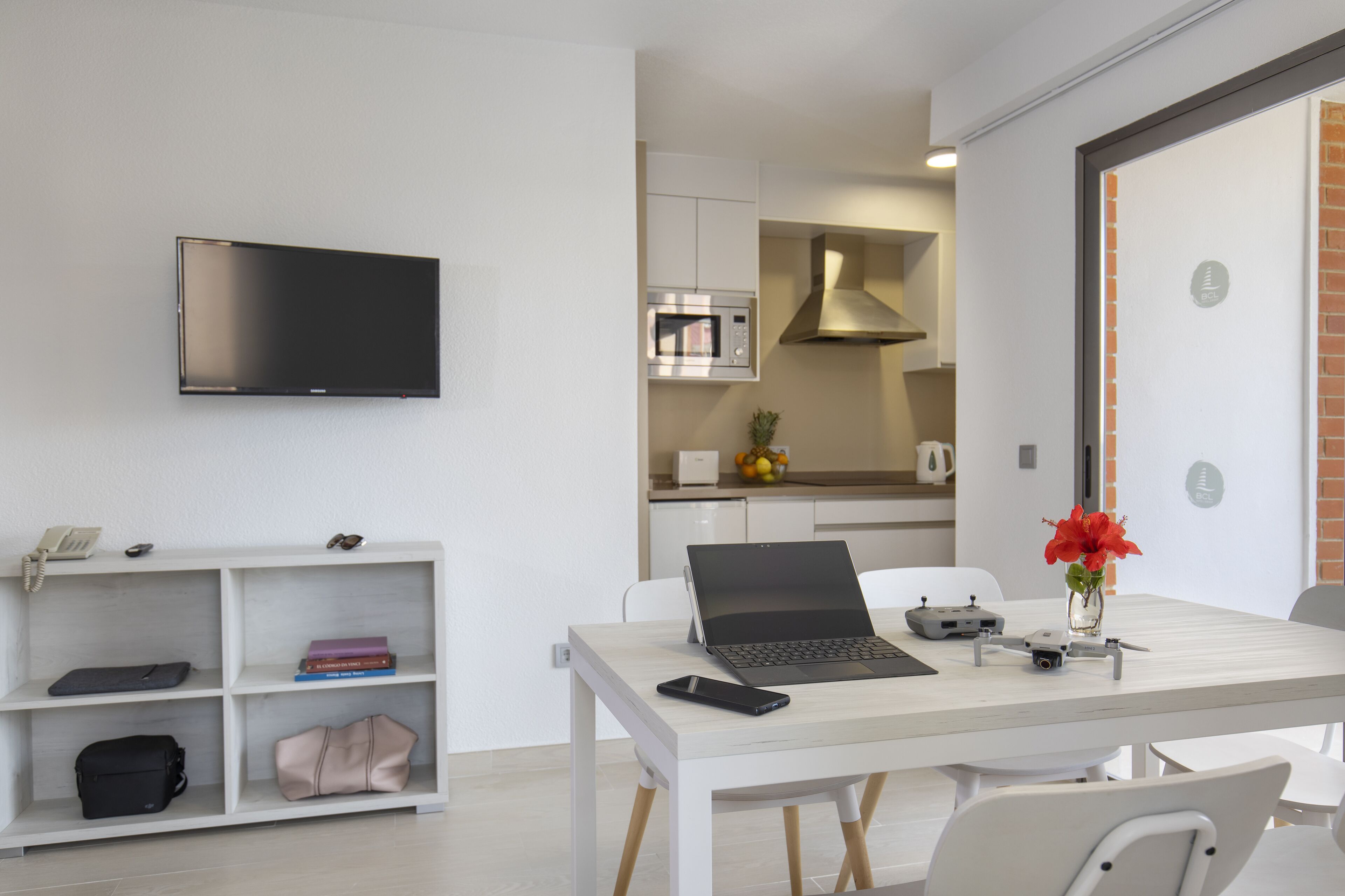Ein modernes, helles Zimmer mit einem an der Wand befestigten Flachbildfernseher, einem Laptop auf einem weißen Tisch, einem Smartphone, einem Spielcontroller und einer Vase mit Blumen.