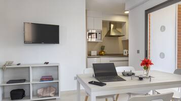 Ein modernes, helles Zimmer mit einem an der Wand befestigten Flachbildfernseher, einem Laptop auf einem weißen Tisch, einem Smartphone, einem Spielcontroller und einer Vase mit Blumen.